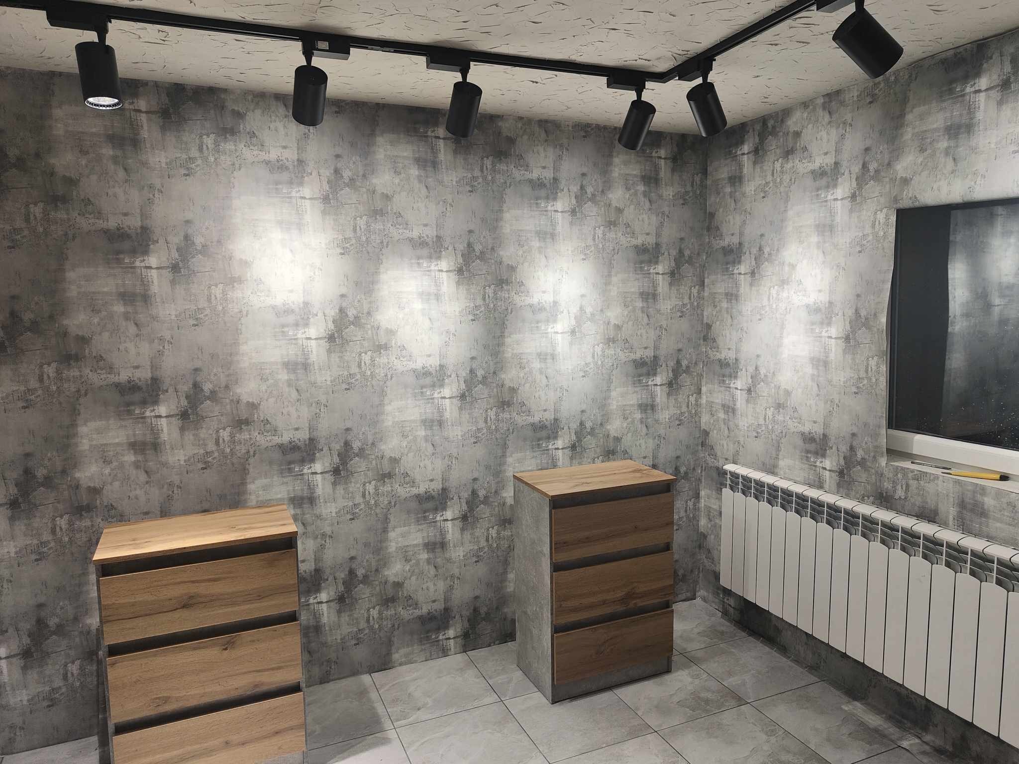 Wykończone wnętrze pokoju z szarą ścianą o nieregularnej fakturze, oświetleniem szynowym i dwiema drewnianymi komodami na tle grzejnika.