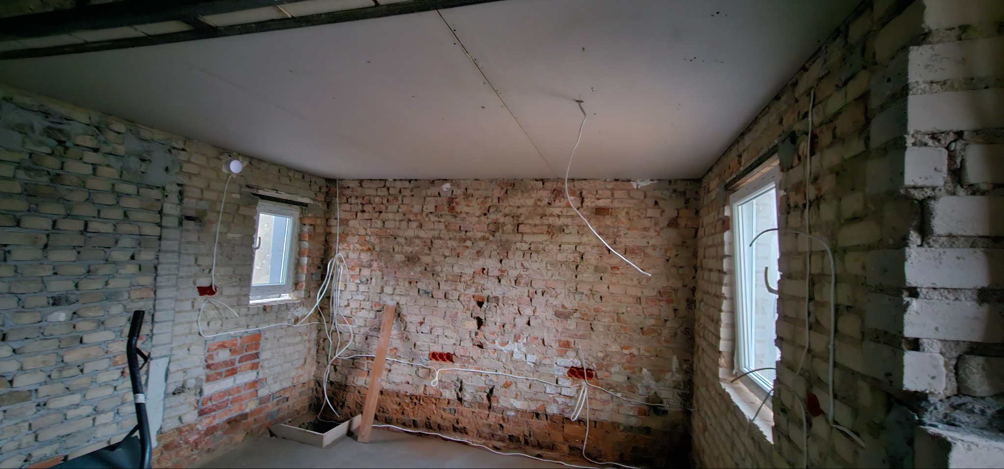 Pokój w trakcie remontu: surowe ceglane ściany z widoczną instalacją elektryczną, częściowo zamontowana płyta karton-gips na suficie, okna. Wnętrze w stanie deweloperskim.