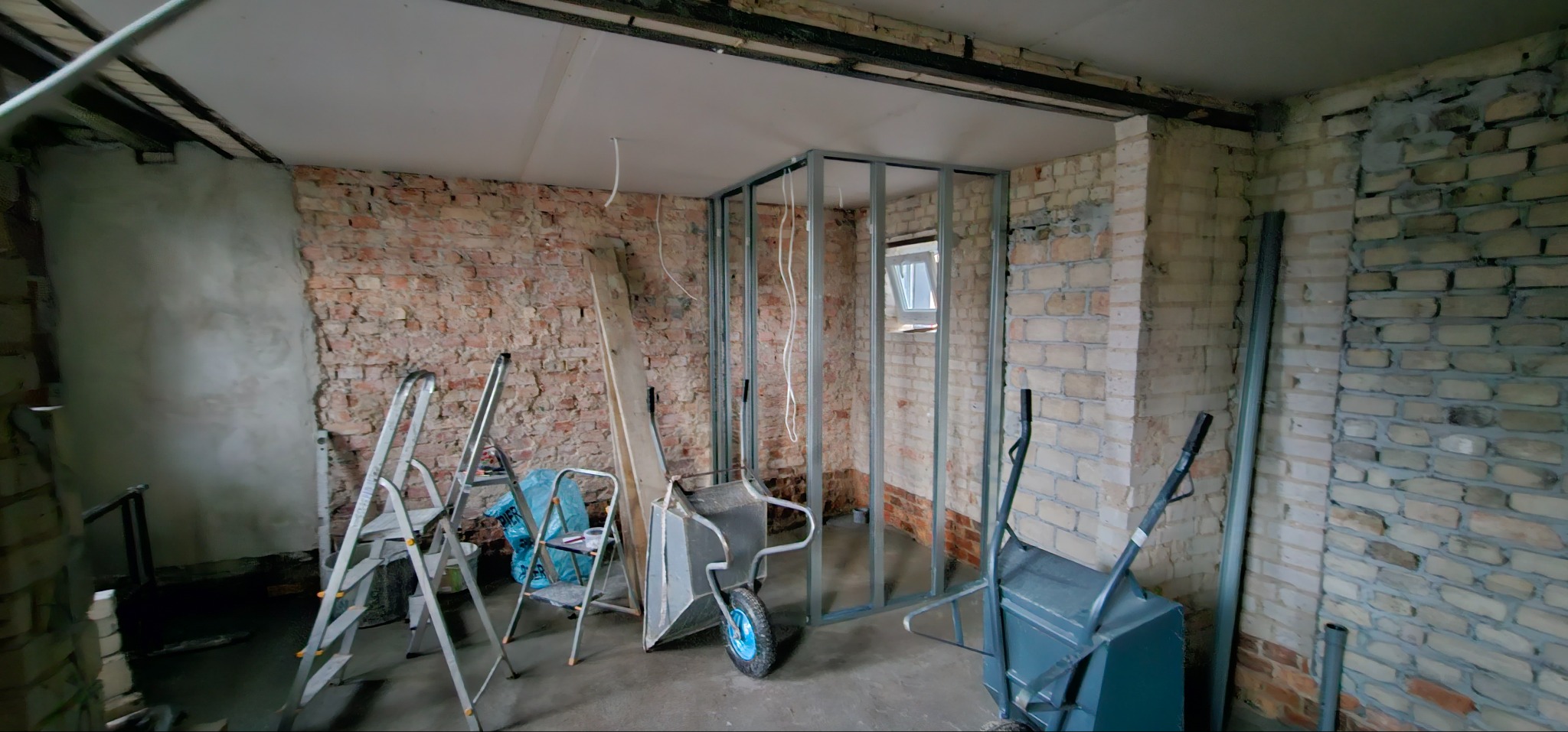 Wnętrze w trakcie remontu: surowa cegła, metalowa konstrukcja ścianki działowej, drabiny, taczki. Widoczne etapy prac wykończeniowych.