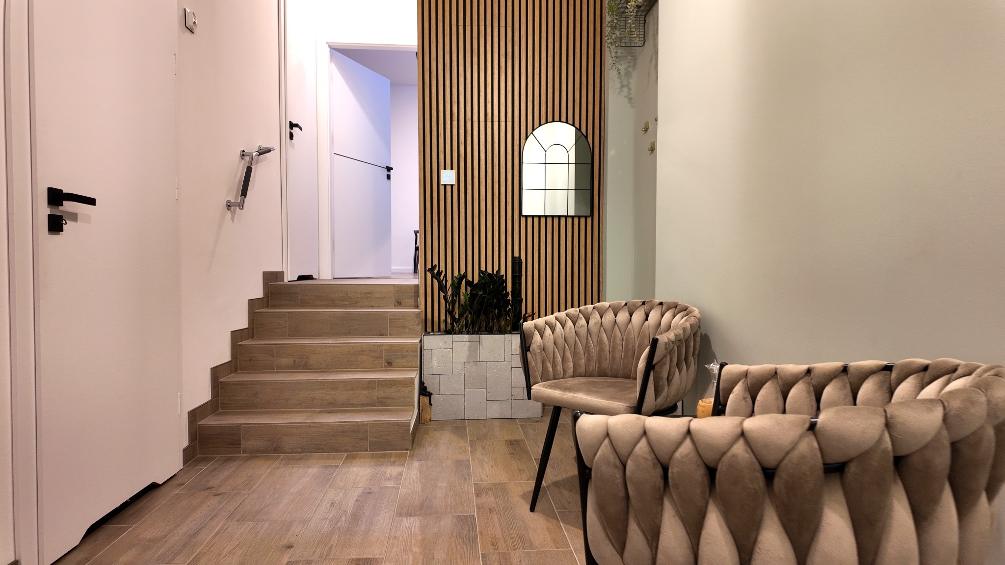 Nowoczesne wnętrze gabinetu z beżowymi fotelami, drewnianą ścianą z lustrem i schodami. Stonowana kolorystyka, minimalistyczny design, przytulna atmosfera.