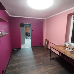 Karol Krankowski - Pokój po malowaniu ścian na różowo i fioletowo, z drewnianą podłogą i widocznymi półkami. W tle otwarte drzwi do innego pomieszczenia.