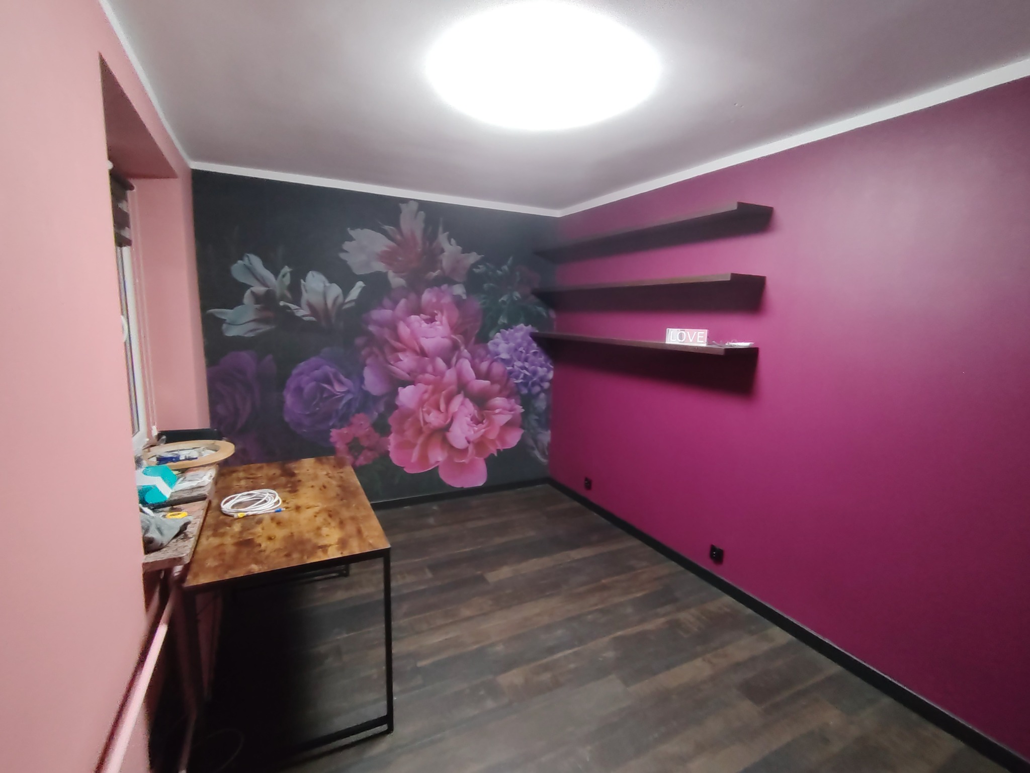 Pokój po odświeżeniu: ciemna podłoga, ściana w kolorze fuksji z półkami, ściana z fototapetą w kwiaty. Na pierwszym planie stół z akcesoriami.