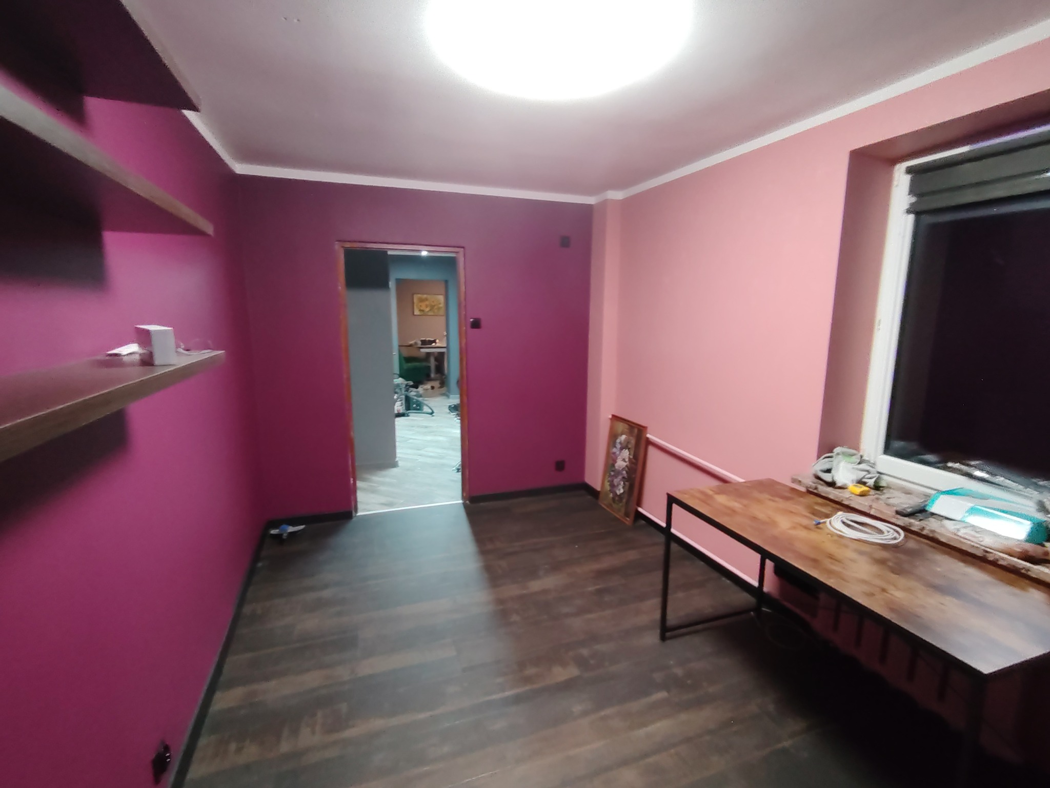 Pokój po malowaniu ścian na różowo i fioletowo, z drewnianą podłogą i widocznymi półkami. W tle otwarte drzwi do innego pomieszczenia.