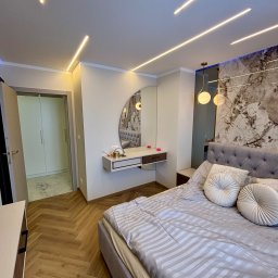 PK-BUD Paweł Kamiński - Nowoczesna sypialnia z parkietem w jodełkę, podświetlanym lustrem nad toaletką i dekoracyjną ścianą z marmurowym wzorem. Oświetlenie liniowe LED na suficie.