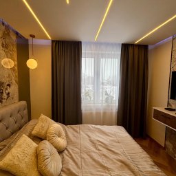 PK-BUD Paweł Kamiński - Nowoczesna sypialnia z podświetlanym sufitem, marmurową ścianą i beżowymi zasłonami. Widoczne łóżko z dekoracyjnymi poduszkami i lampa wisząca.