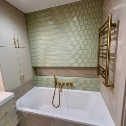 PK-BUD Paweł Kamiński - Nowoczesna łazienka z wanną, zieloną mozaiką na ścianie i złotą armaturą. Minimalistyczny design, ciepłe oświetlenie, szafki w zabudowie. Eleganckie wykończenie.