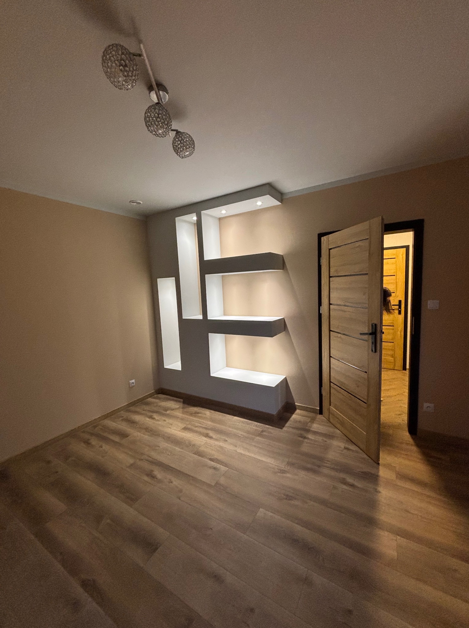 Wykończone wnętrze pokoju z drewnianą podłogą, otwartymi drzwiami i dekoracyjną, podświetlaną półką w ścianie. Ściany w odcieniach beżu i szarości. Nowoczesna lampa sufitowa.