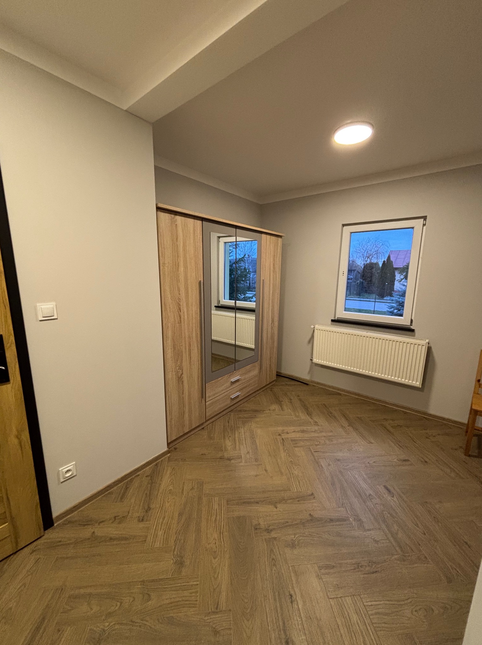 Jasne wnętrze pokoju z drewnianą podłogą w jodełkę, szafą z lustrem i oknem z grzejnikiem. Świeże, neutralne kolory ścian i sufitu.