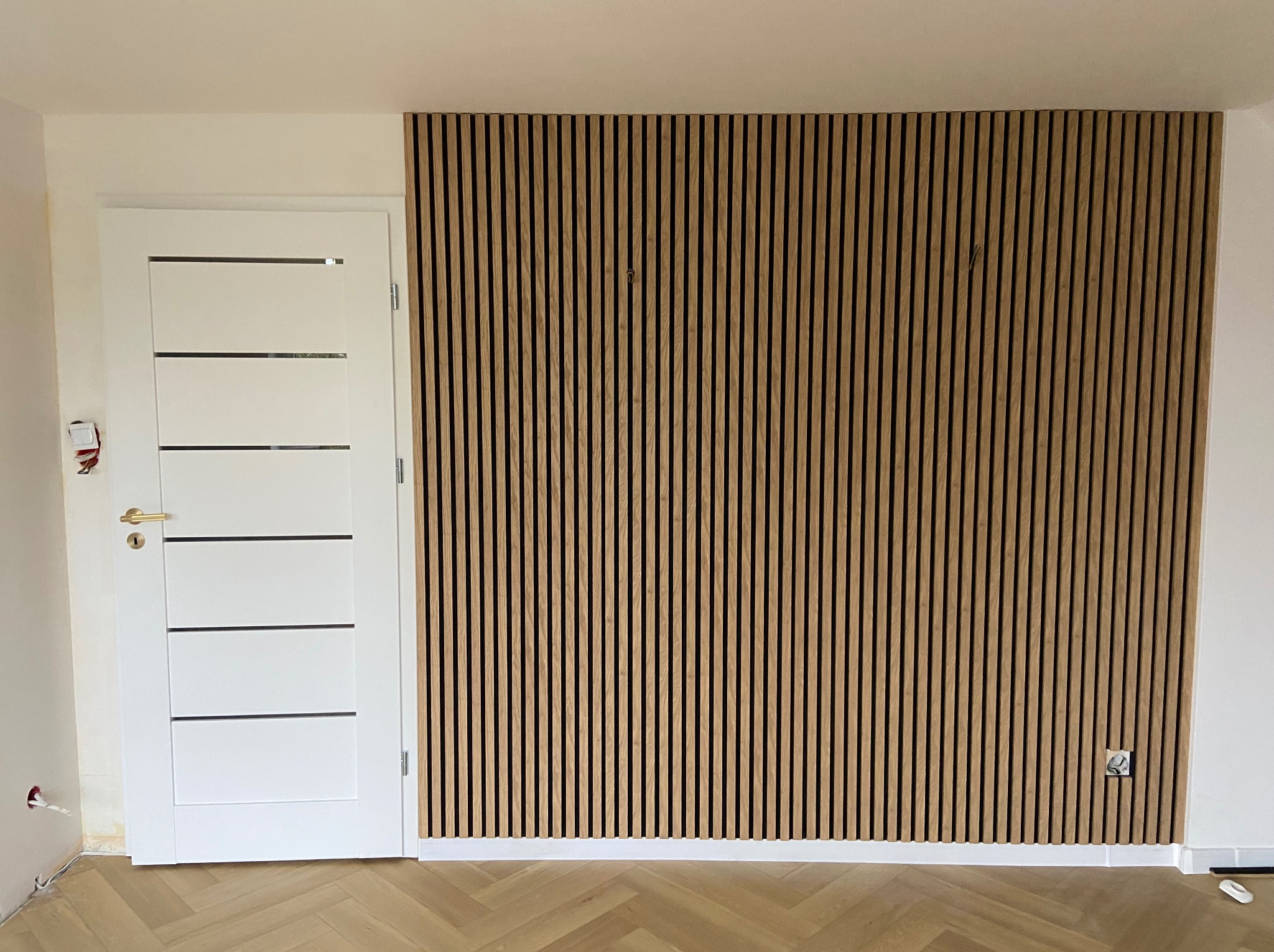 Nowoczesny panel ścienny z drewnianych listew w jasnym odcieniu, obok białe drzwi z poziomymi przeszkleniami. Parkiet w jodełkę na podłodze. Widoczne gniazdko elektryczne.