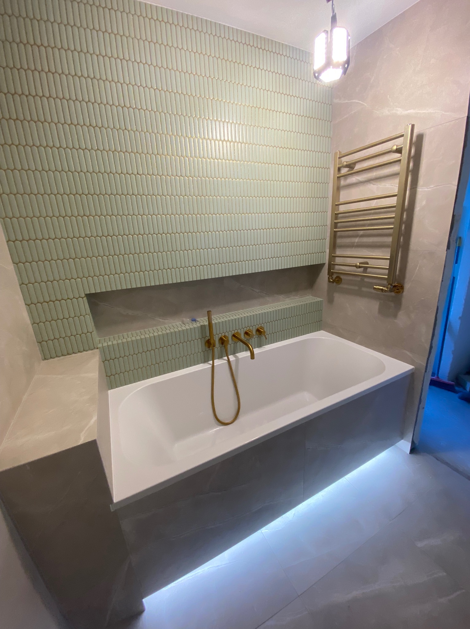 Wykończona łazienka z wanną podświetloną LED, złota armatura i grzejnik drabinkowy. Ściana wyłożona oryginalną, pistacjową mozaiką. Nowoczesny design.