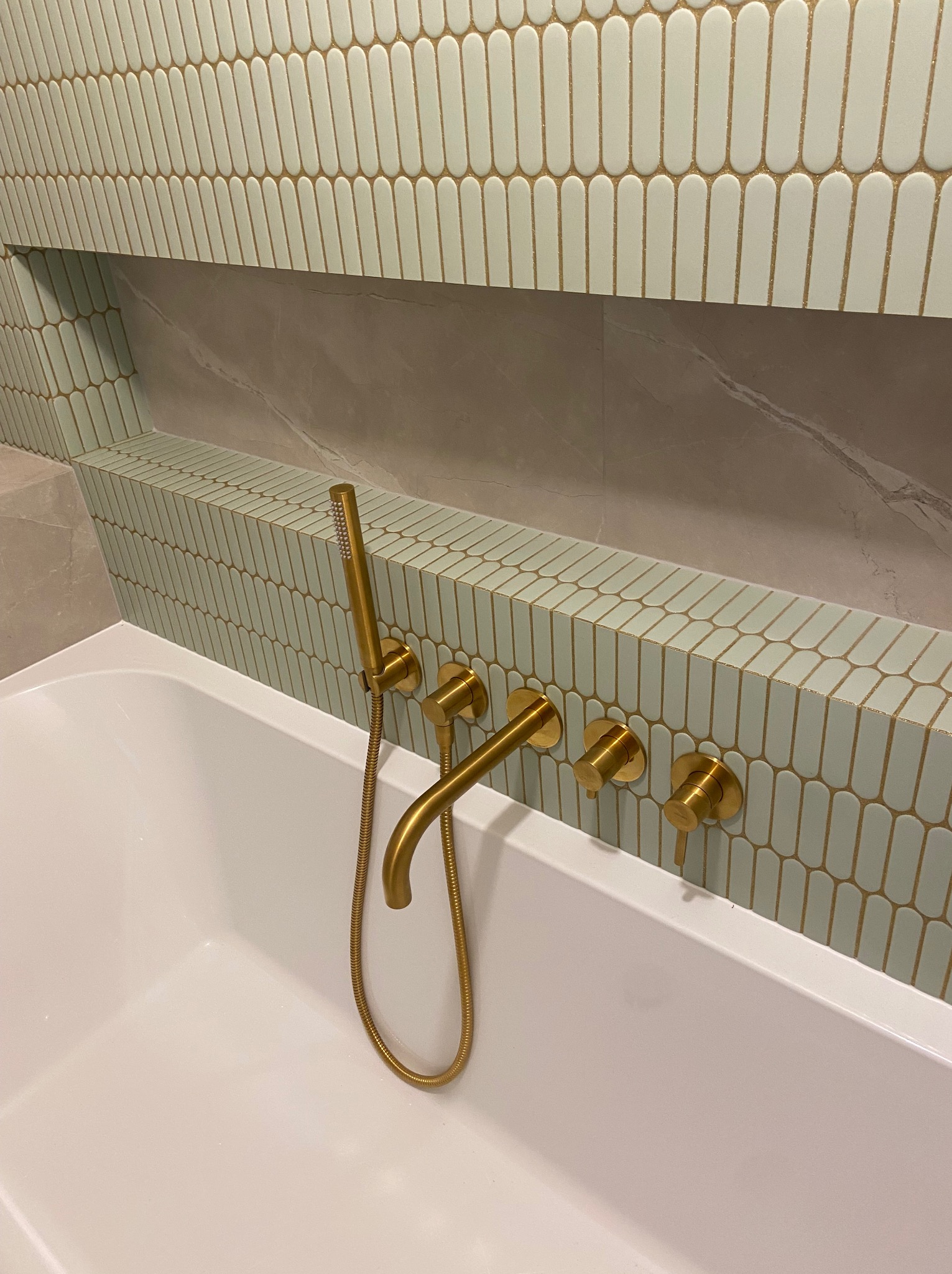 Biała wanna z baterią w kolorze złotym i dekoracyjną ścianą z płytek w kształcie pastylek w łazience. Widoczna wnęka w ścianie wyłożona płytkami imitującymi marmur.