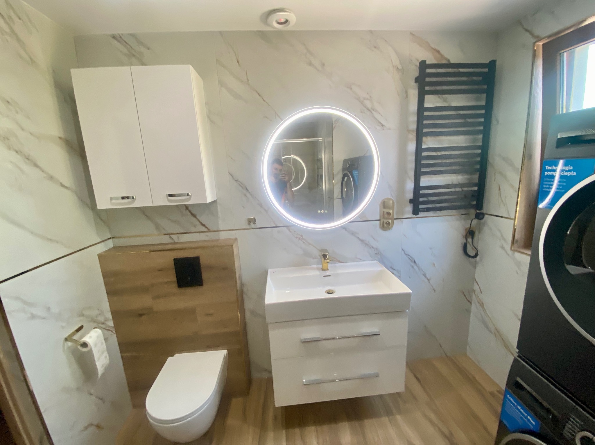Nowoczesna łazienka z marmurowymi płytkami, okrągłym lustrem z podświetleniem LED, wiszącą szafką z umywalką i grzejnikiem drabinkowym. Widoczna toaleta i pralka.