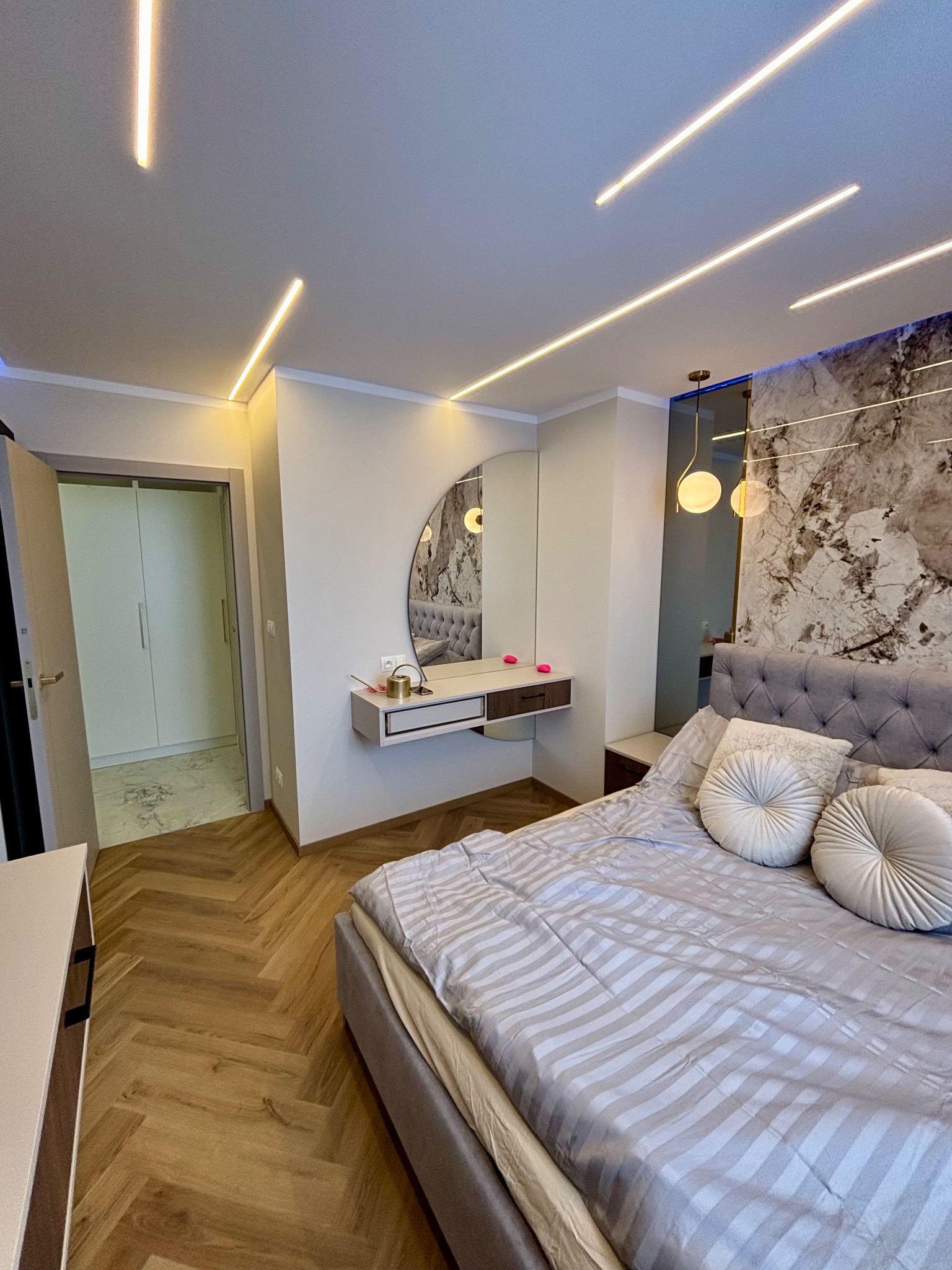 Nowoczesna sypialnia z parkietem w jodełkę, podświetlanym lustrem nad toaletką i dekoracyjną ścianą z marmurowym wzorem. Oświetlenie liniowe LED na suficie.