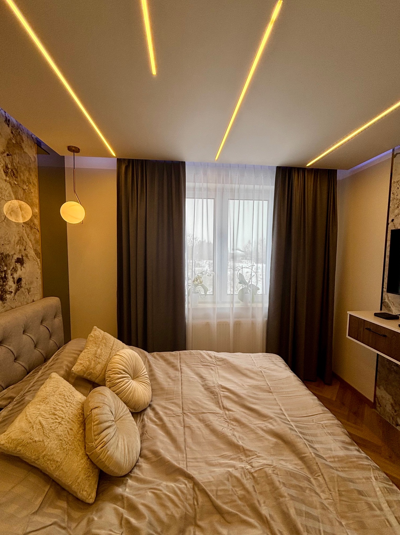 Nowoczesna sypialnia z podświetlanym sufitem, marmurową ścianą i beżowymi zasłonami. Widoczne łóżko z dekoracyjnymi poduszkami i lampa wisząca.