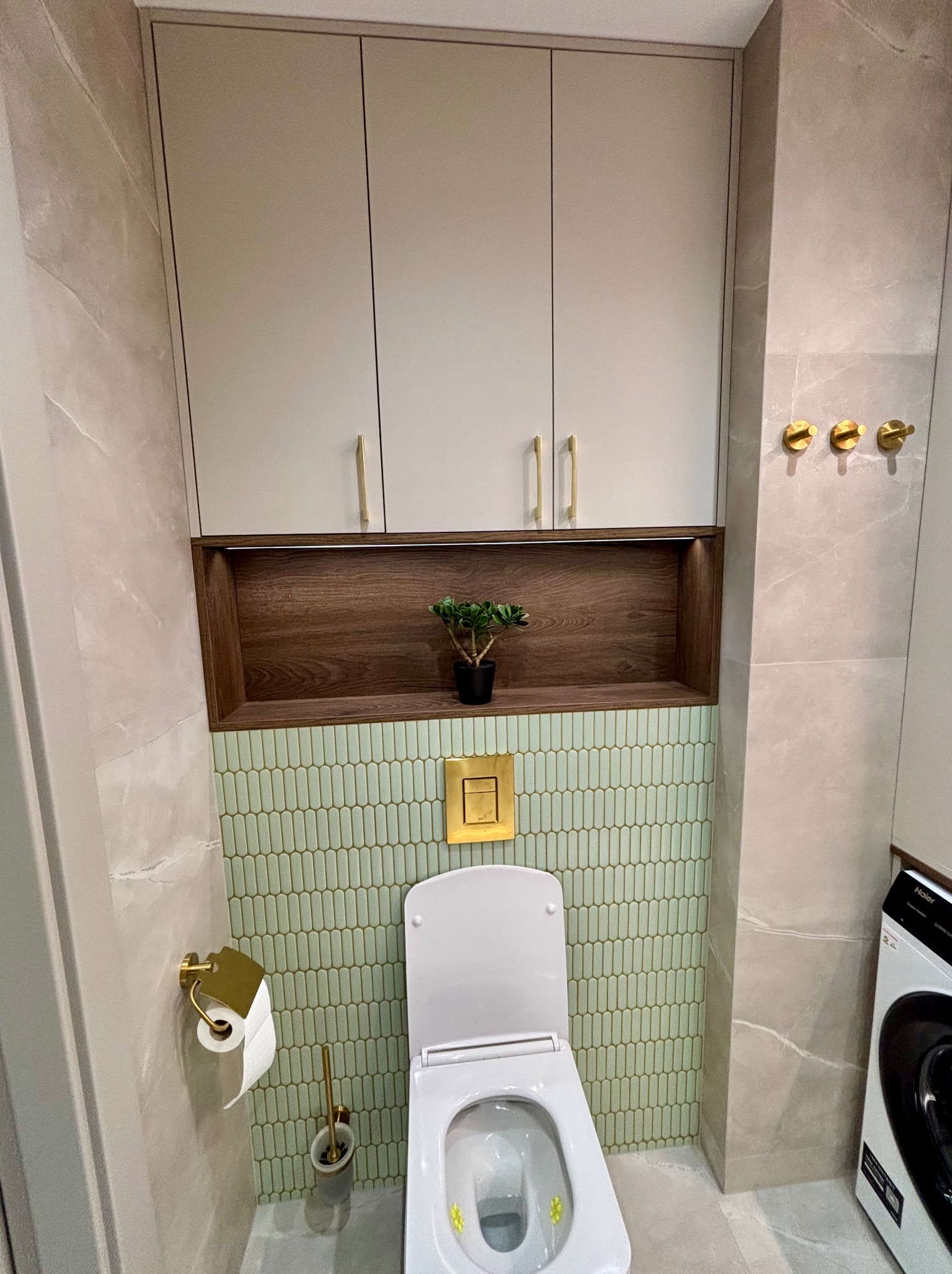 Nowoczesna łazienka po remoncie: beżowe płytki imitujące marmur, zielona mozaika, wisząca toaleta z złotymi akcentami i szafka z drewnianą wnęką.