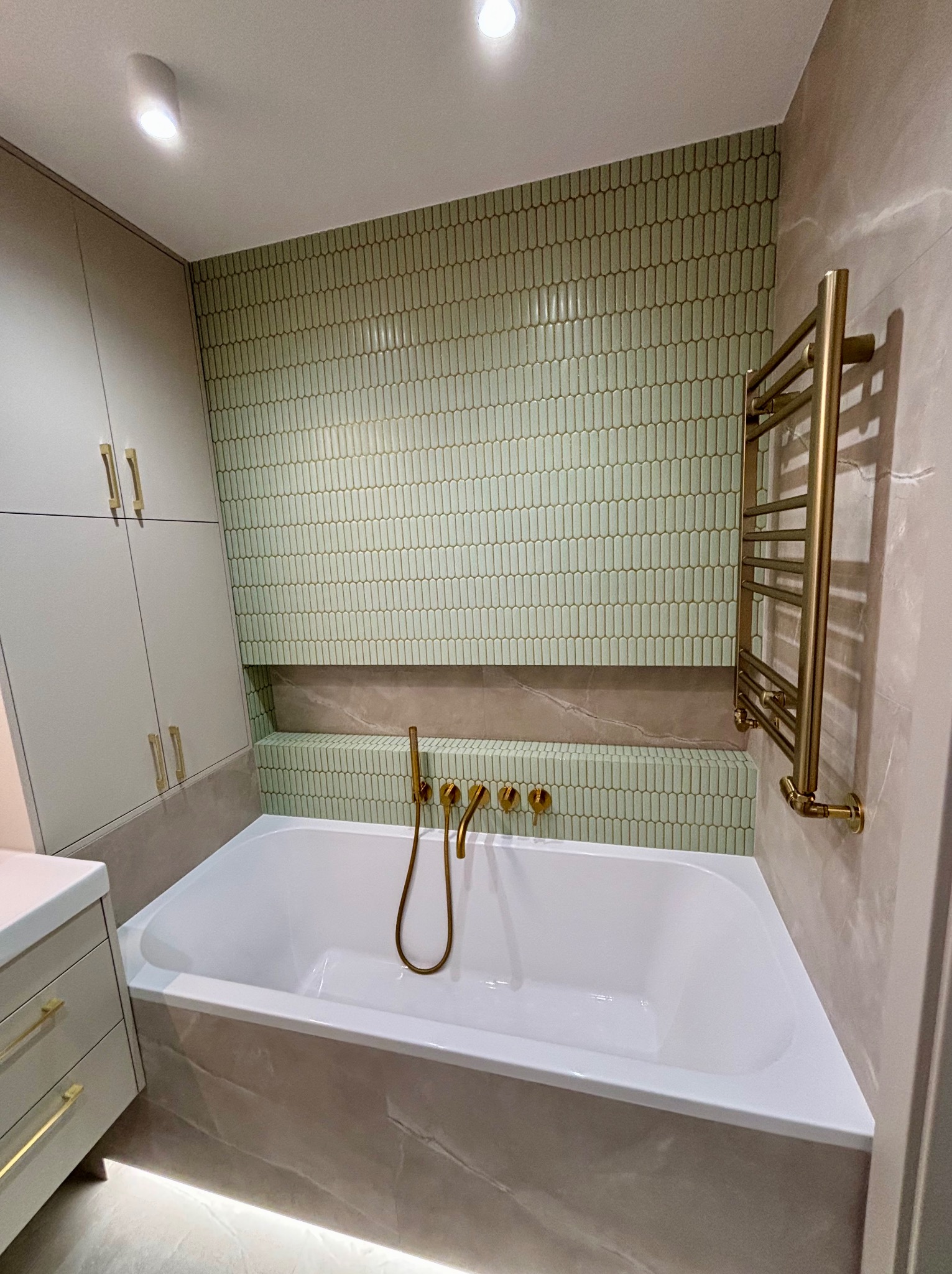 Nowoczesna łazienka z wanną, zieloną mozaiką na ścianie i złotą armaturą. Minimalistyczny design, ciepłe oświetlenie, szafki w zabudowie. Eleganckie wykończenie.