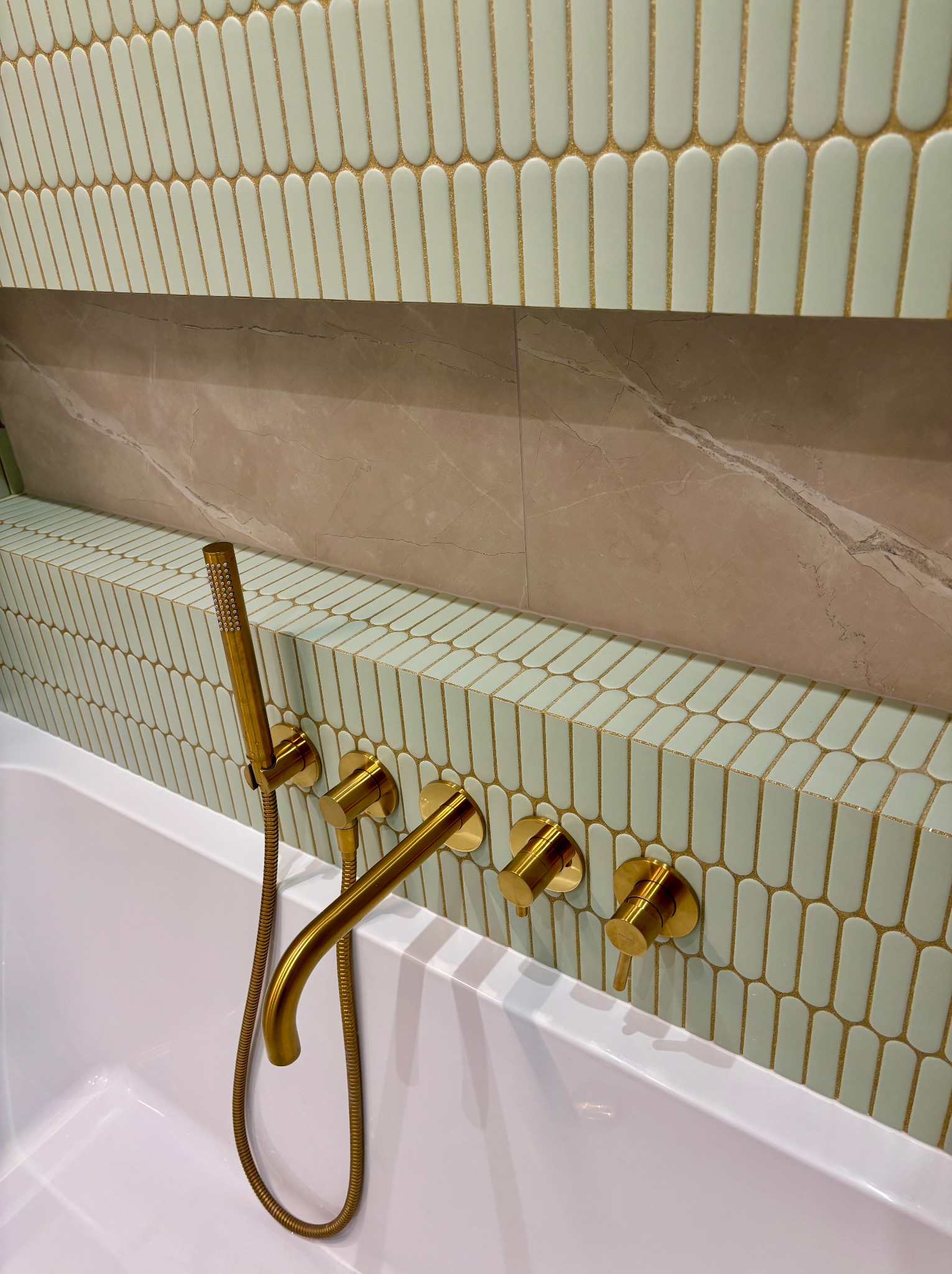 Biała wanna z baterią w kolorze złota na tle ściany wyłożonej podłużnymi, pastelowymi płytkami z dekoracyjną złotą fugą i fragmentem ściany z imitacją marmuru.