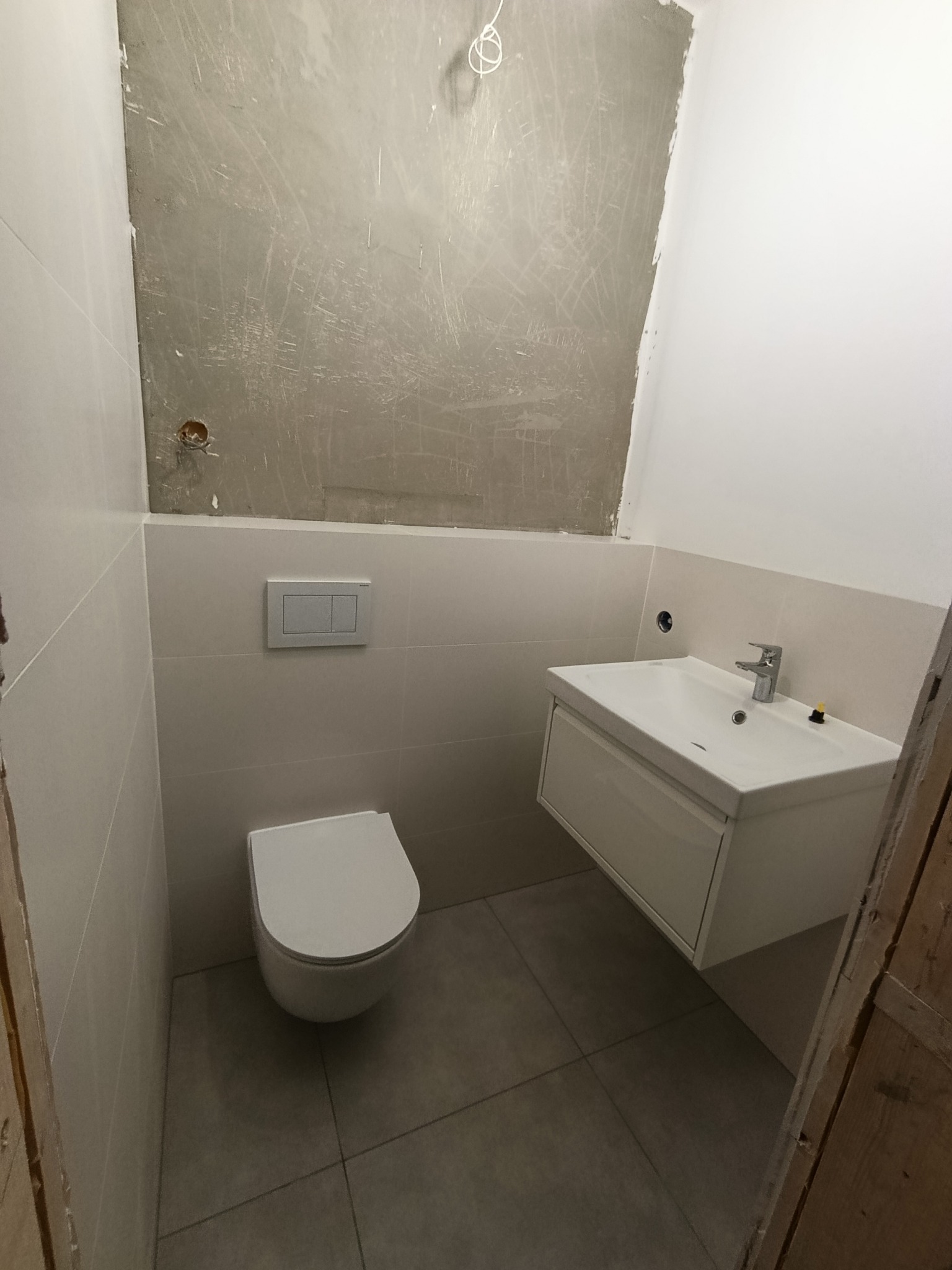 Wnętrze łazienki w trakcie remontu: częściowo wykończone płytki, zamontowana toaleta i umywalka z szafką, surowa ściana w górnej części. Widoczne niedokończone prace.