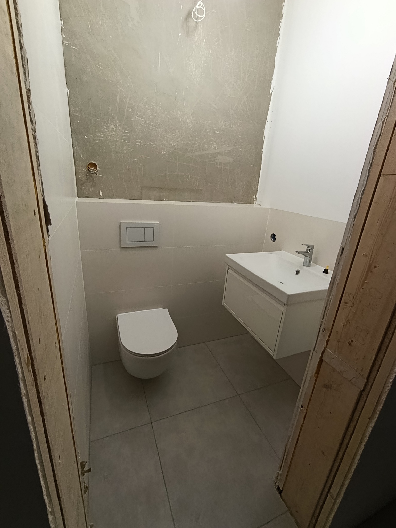 Wnętrze łazienki w trakcie remontu: biała umywalka z szafką, podwieszana toaleta, szare płytki podłogowe. Ściana częściowo przygotowana do malowania.