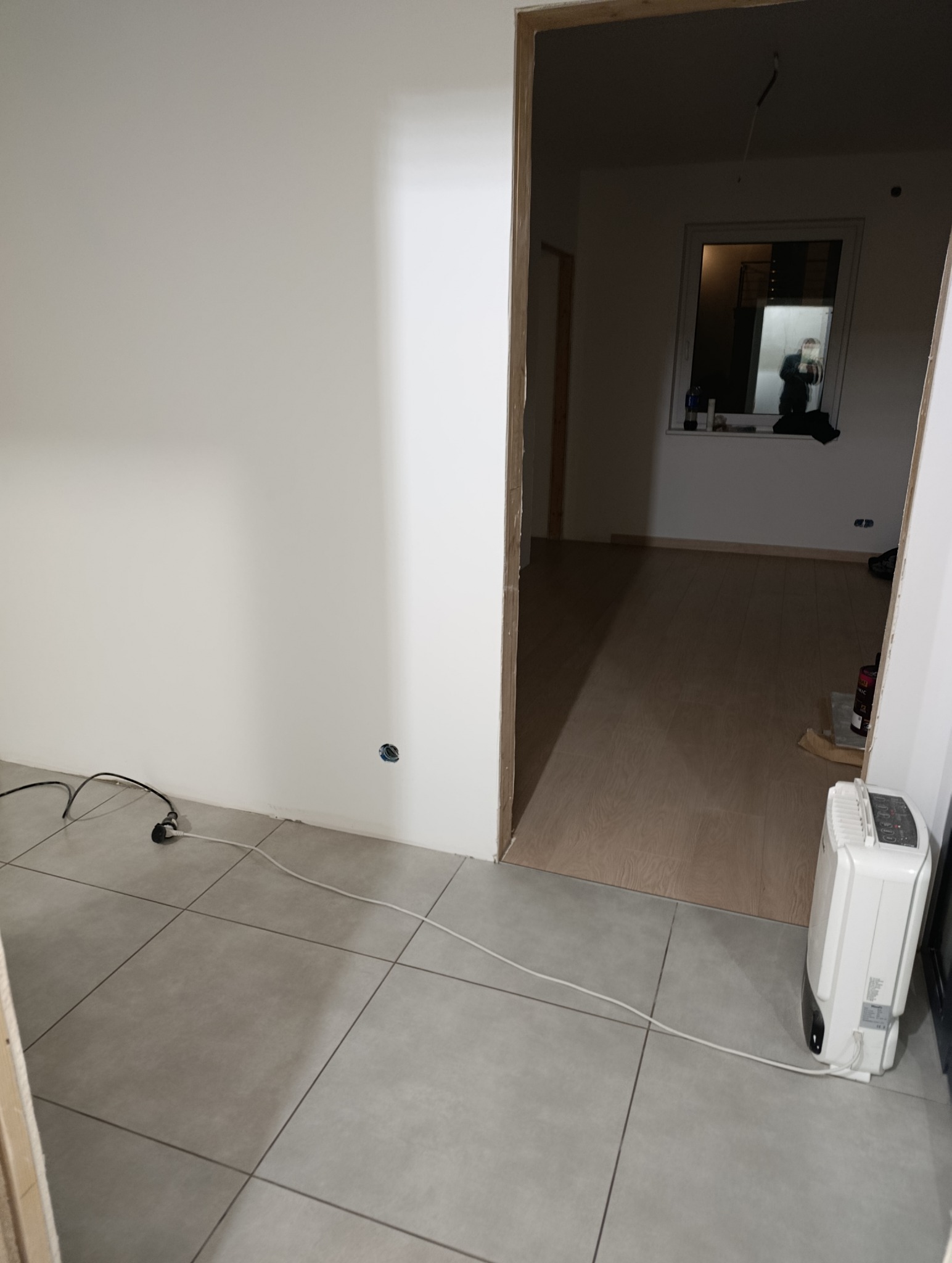 Wnętrze w trakcie remontu: widoczne nowe panele podłogowe, świeżo pomalowane ściany i przygotowane ościeżnice drzwiowe. W tle okno z odbiciem osoby.