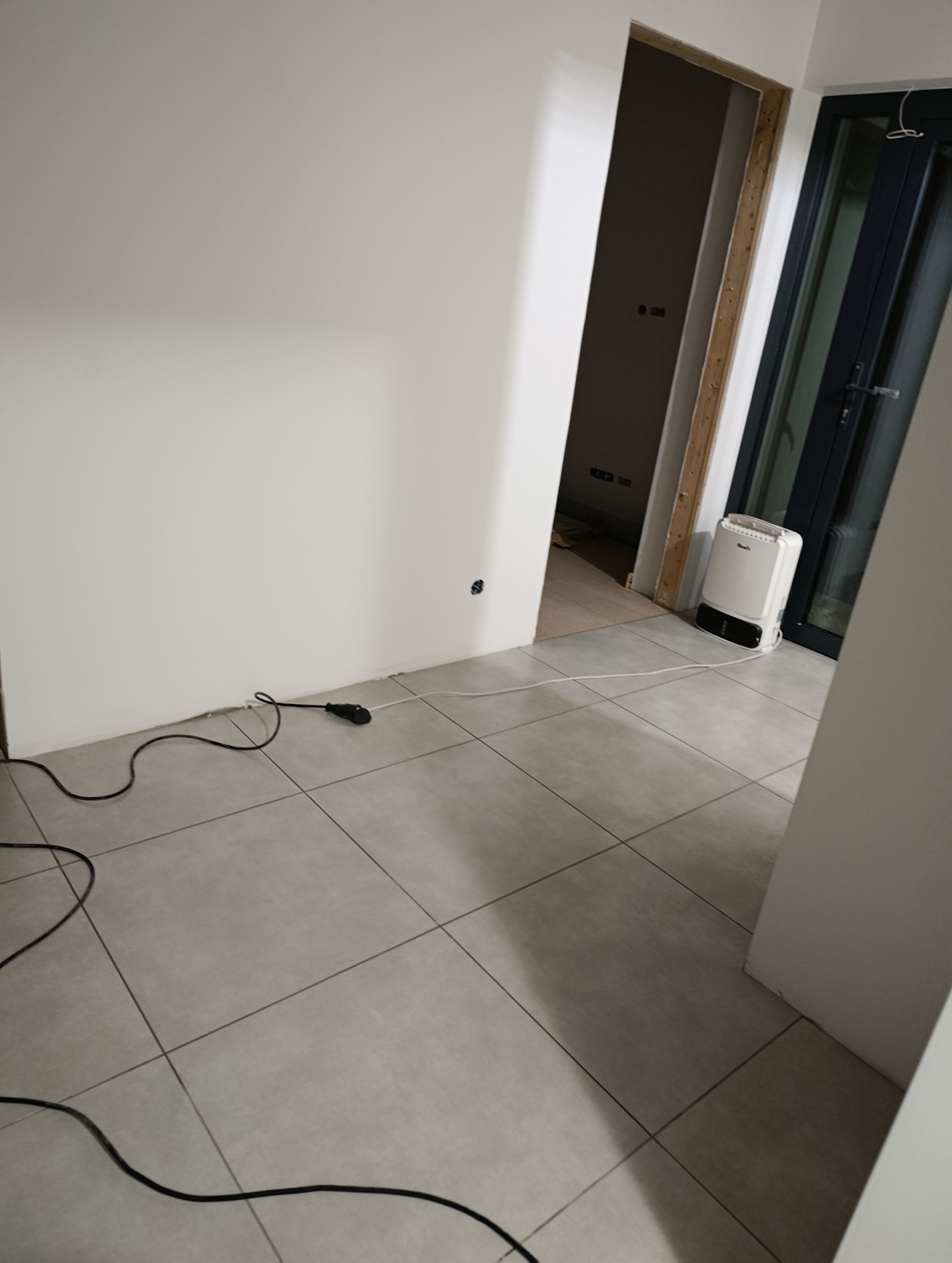Szare płytki podłogowe w trakcie remontu, widoczne kable i grzejnik elektryczny. Otwór drzwiowy bez ościeżnicy, białe ściany. Ujęcie z góry, perspektywa ukośna.