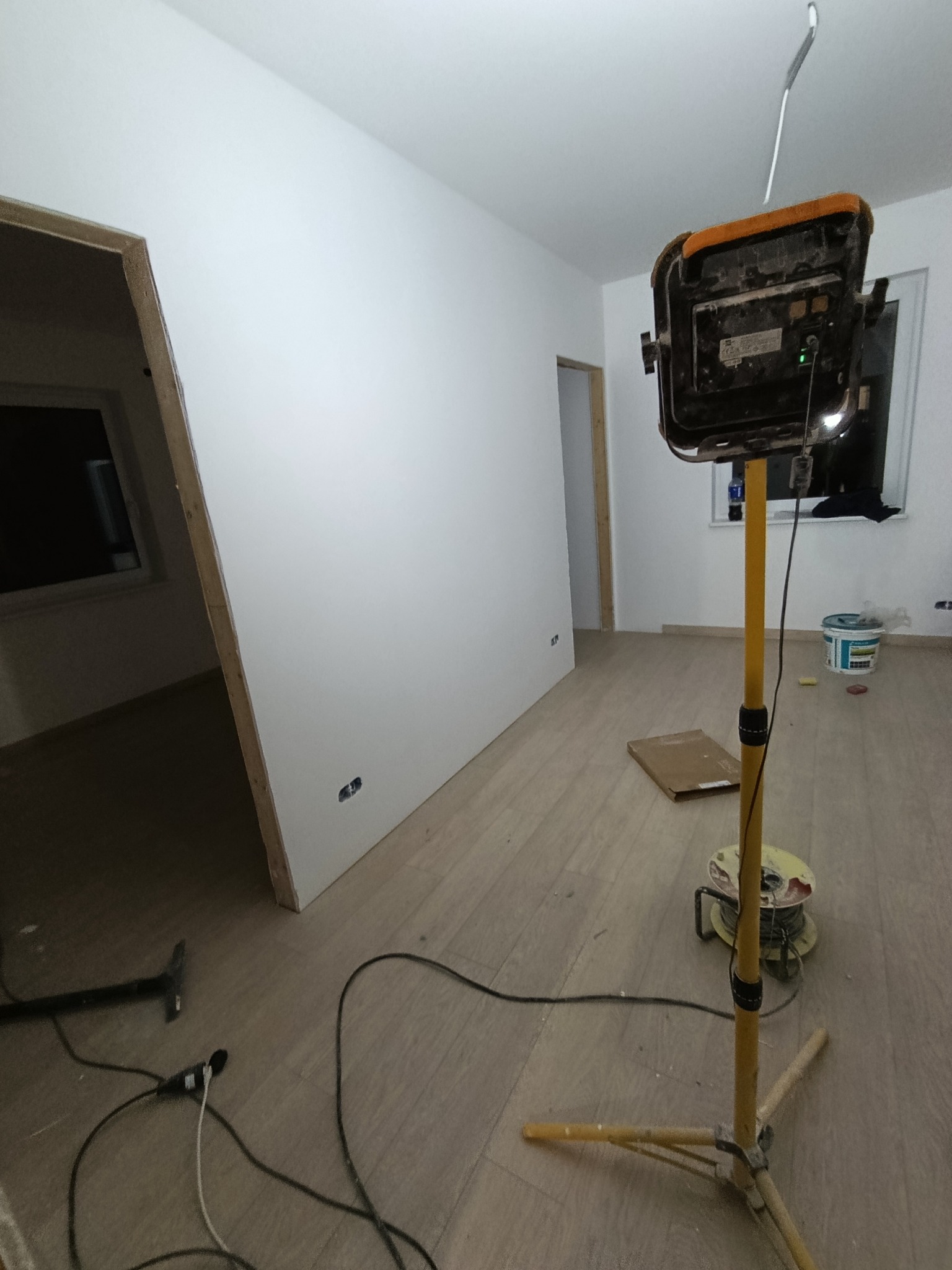Wnętrze w trakcie prac wykończeniowych: białe ściany, drewnopodobna podłoga, widoczna lampa budowlana na statywie i narzędzia. Częściowo widoczne ościeżnice drzwi.