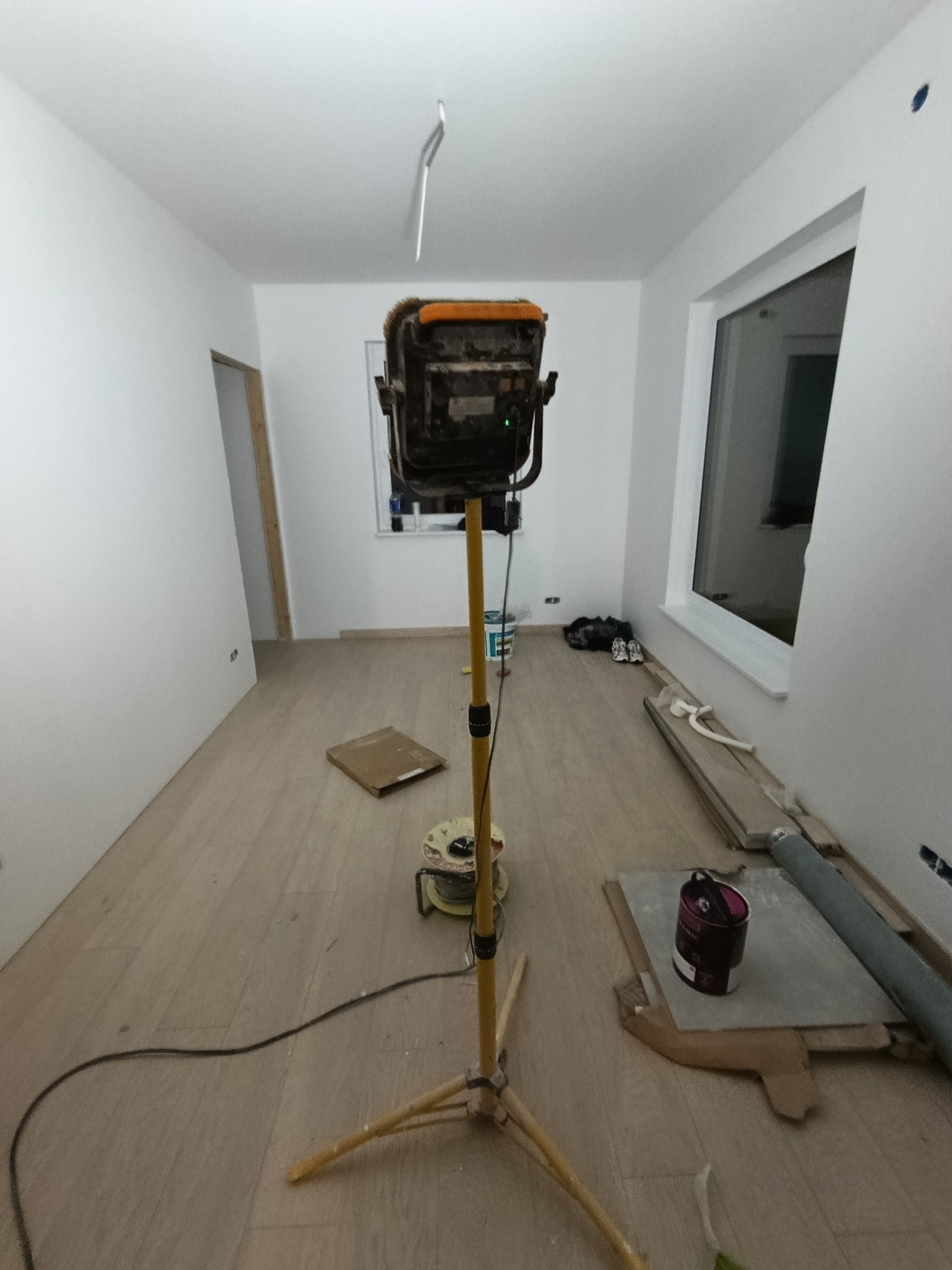 Wnętrze w trakcie remontu: podłoga z paneli, ściany pomalowane na biało, lampa budowlana na statywie, puszka z farbą i materiały budowlane. Widoczne niedoskonałości i etap prac.