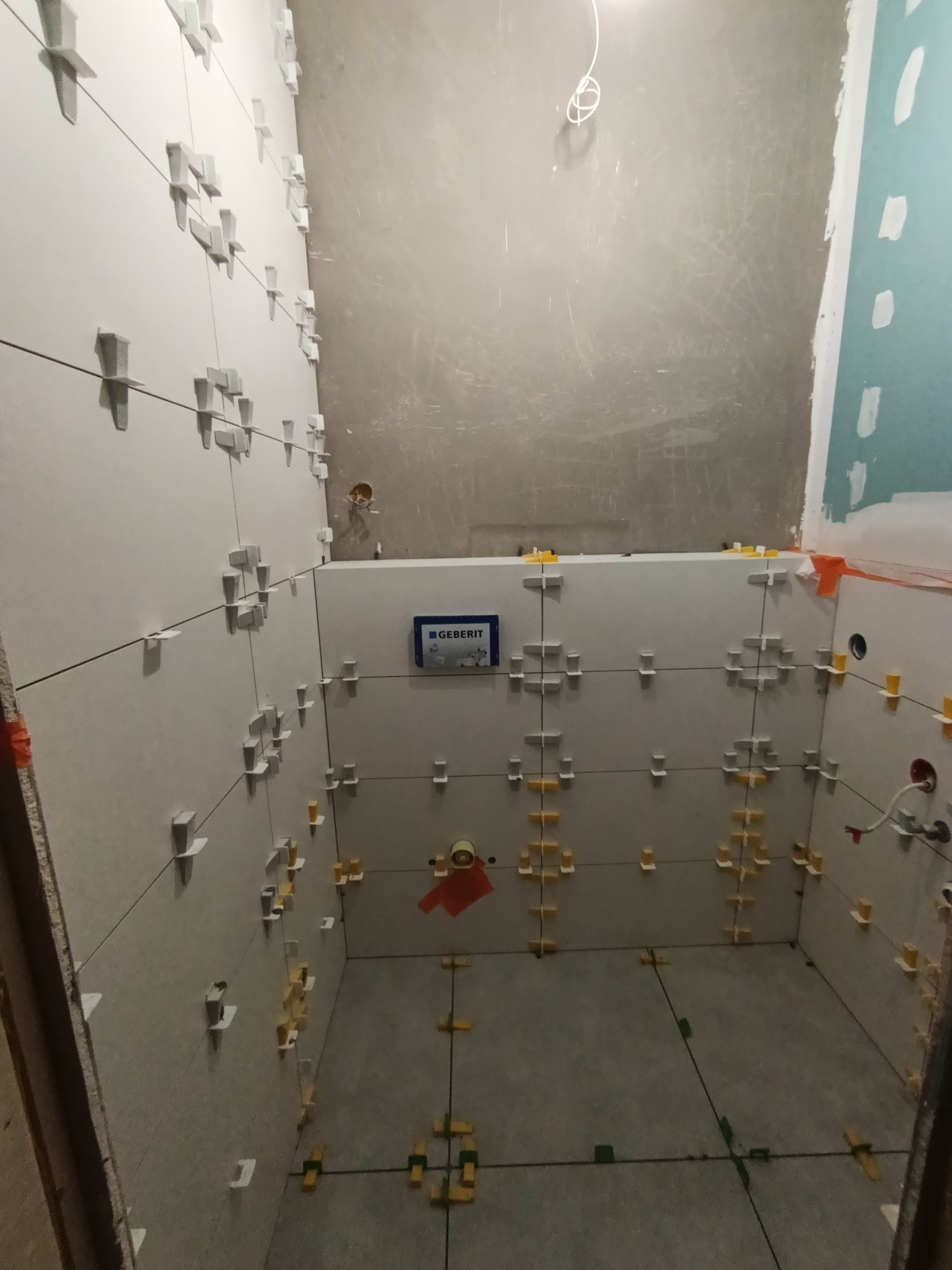 Świeżo ułożone, szare płytki w łazience, z krzyżykami dystansowymi. Widoczna instalacja Geberit. Surowe ściany czekają na wykończenie. Ujęcie z dołu, podkreślające przestrzeń.
