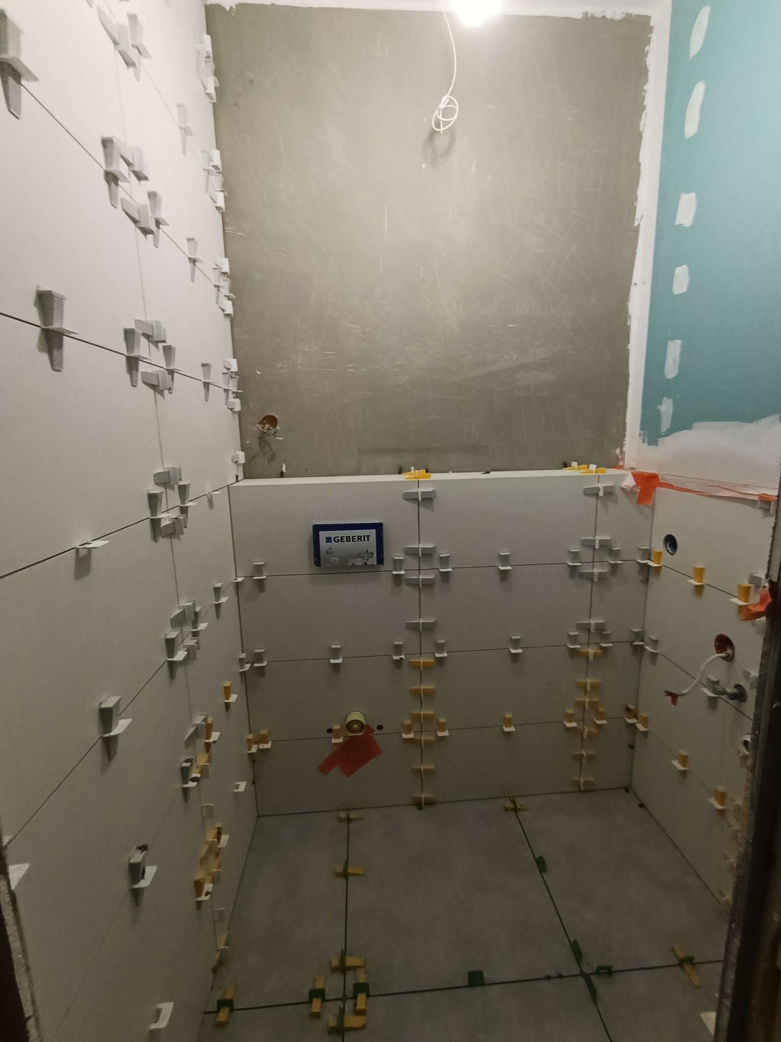 Wnętrze łazienki w trakcie układania płytek, widoczne krzyżyki dystansowe i poziomica laserowa. Częściowo wykończone ściany, surowy beton w górnej części ściany.