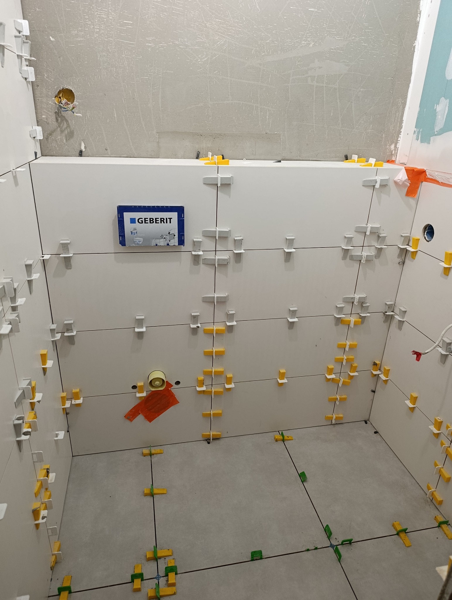 Białe płytki ścienne z systemem poziomowania w trakcie montażu w łazience. Widoczny fragment podłogi z szarych płytek. Na ścianie logo Geberit.