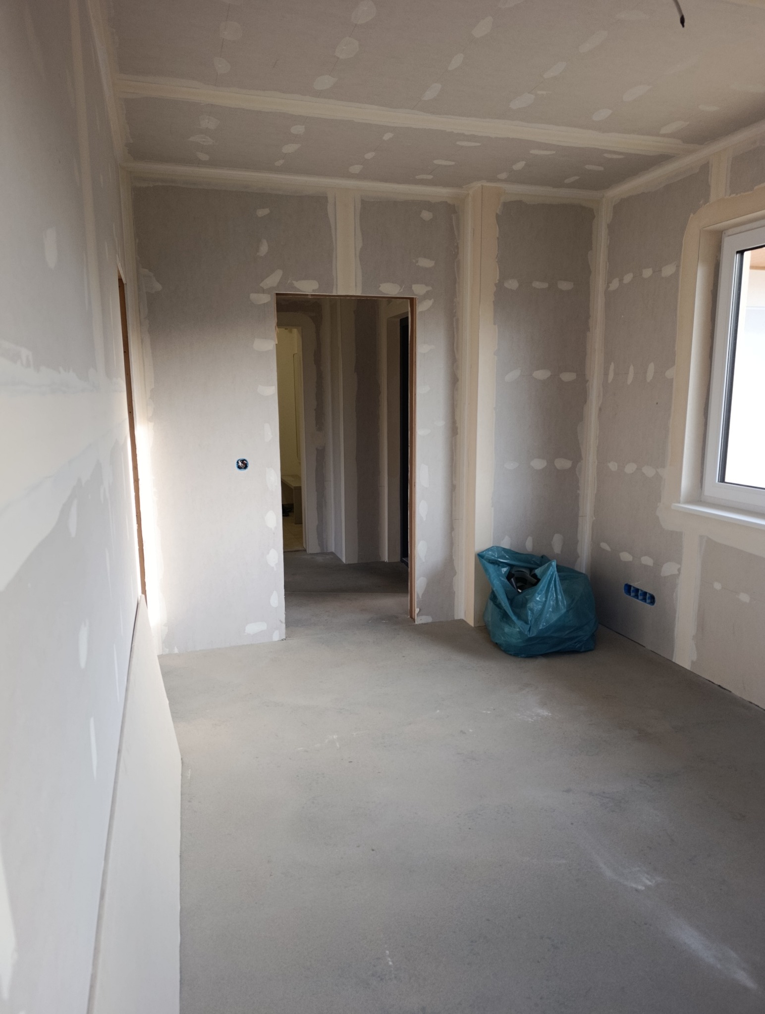 Pokój w trakcie wykańczania: ściany z płyt karton-gips, widoczne łączenia szpachlowane, surowa podłoga, okno i worek z odpadami budowlanymi. Wnętrze w stanie surowym.