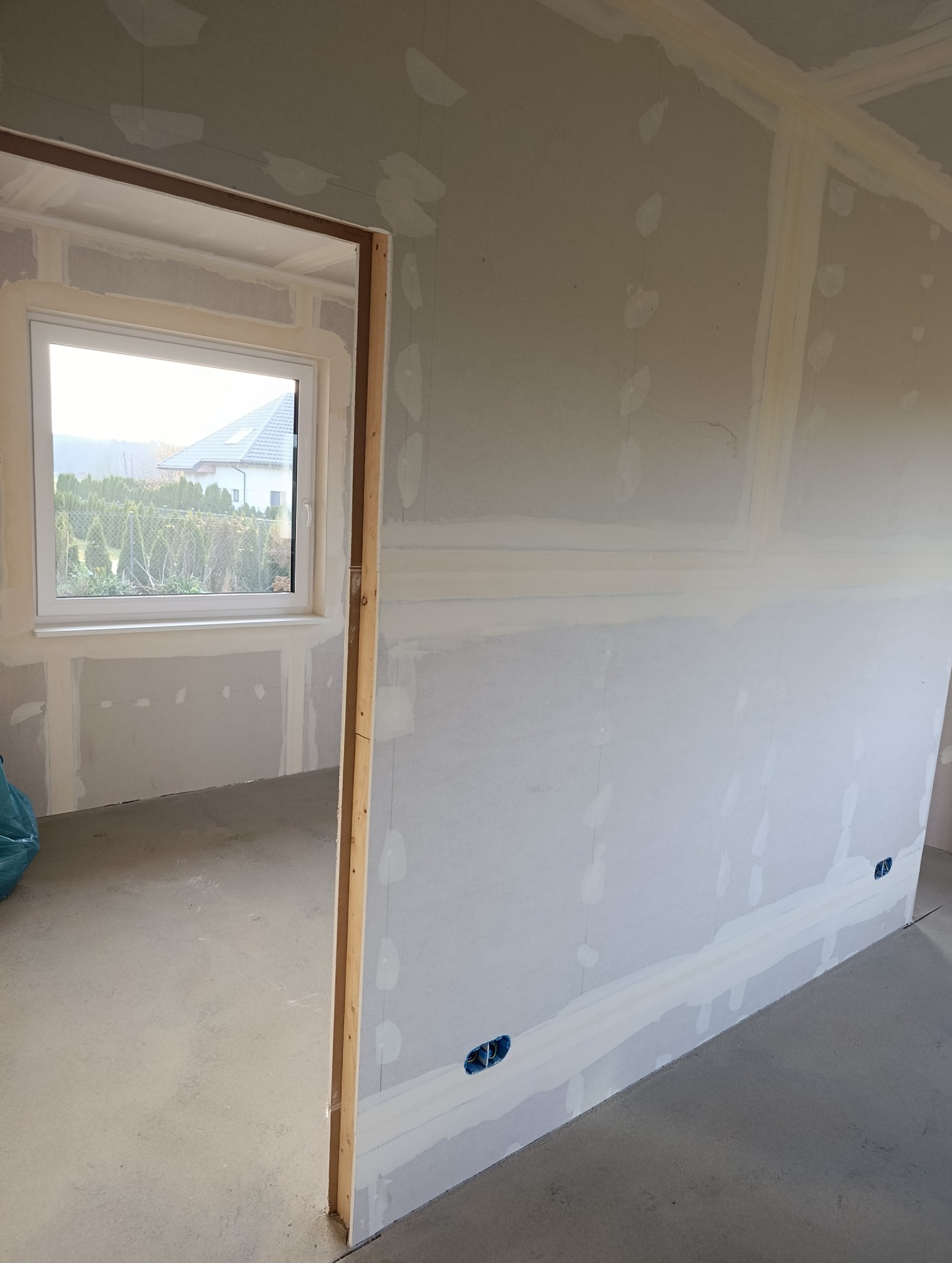 Ściana z płyt kartonowo-gipsowych w trakcie szpachlowania, widoczne gniazdka elektryczne i fragment okna z widokiem na dom i zieleń. Surowy stan wykończenia wnętrza.