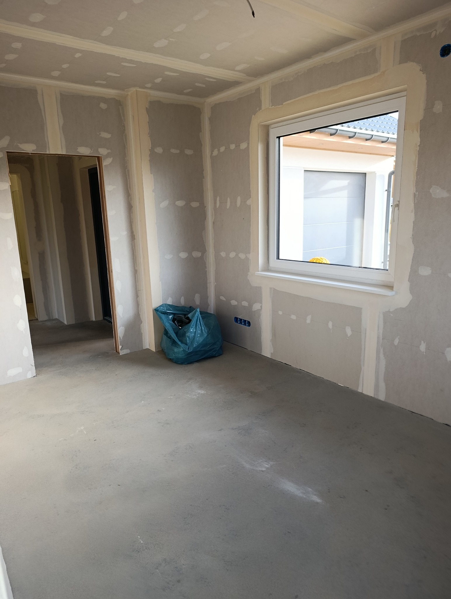 Pokój w trakcie wykańczania: ściany z płyt karton-gips, okno z widokiem na zewnątrz, surowa podłoga, niebieski worek na śmieci. Widoczne puszki elektryczne.