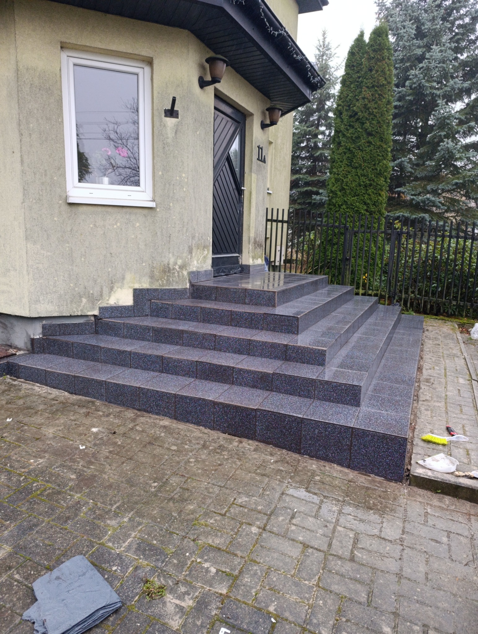 Zewnętrzne schody wejściowe do domu, wyłożone ciemną, błyszczącą terakotą. Widoczne detale wykończenia i fuga. Tło stanowi elewacja budynku i zieleń.