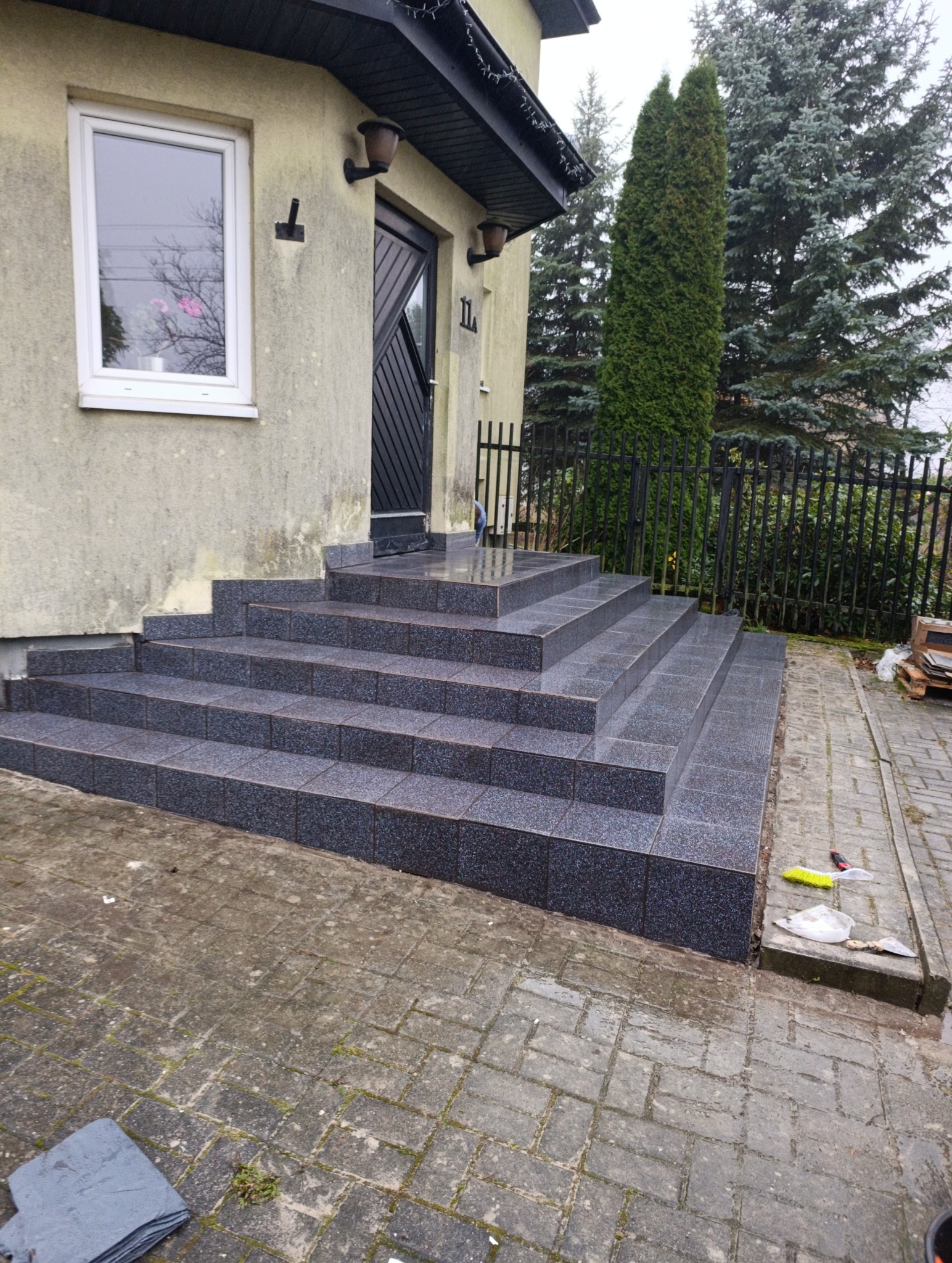 Wykończone płytkami schody wejściowe do domu z czarnymi drzwiami i numerem 11. Widoczne narzędzia i resztki po pracy. Tło: drzewa iglaste i ogrodzenie.