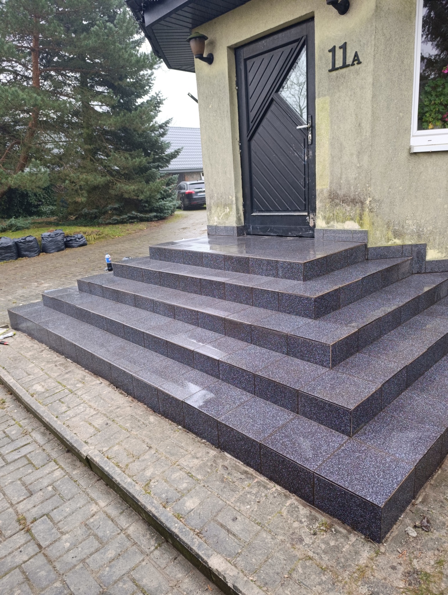 Wykończone płytkami schody wejściowe do domu z czarnymi drzwiami i numerem 11a. Widoczne worki na śmieci i butelka. Pochmurny dzień.