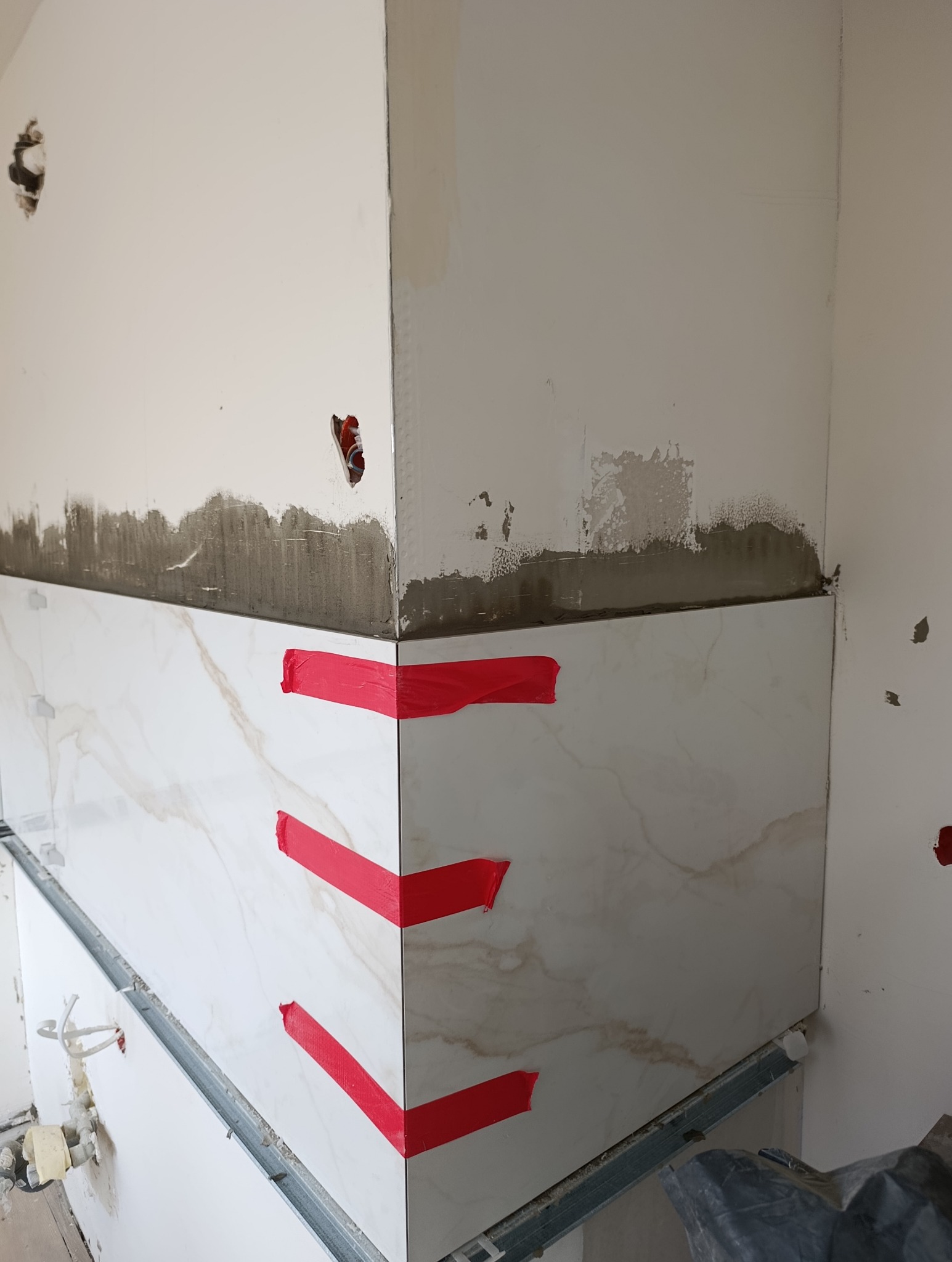 Ściana w trakcie wykańczania z marmurowymi płytkami, zabezpieczonymi czerwoną taśmą. Widoczne ślady po kleju i instalacji elektrycznej.