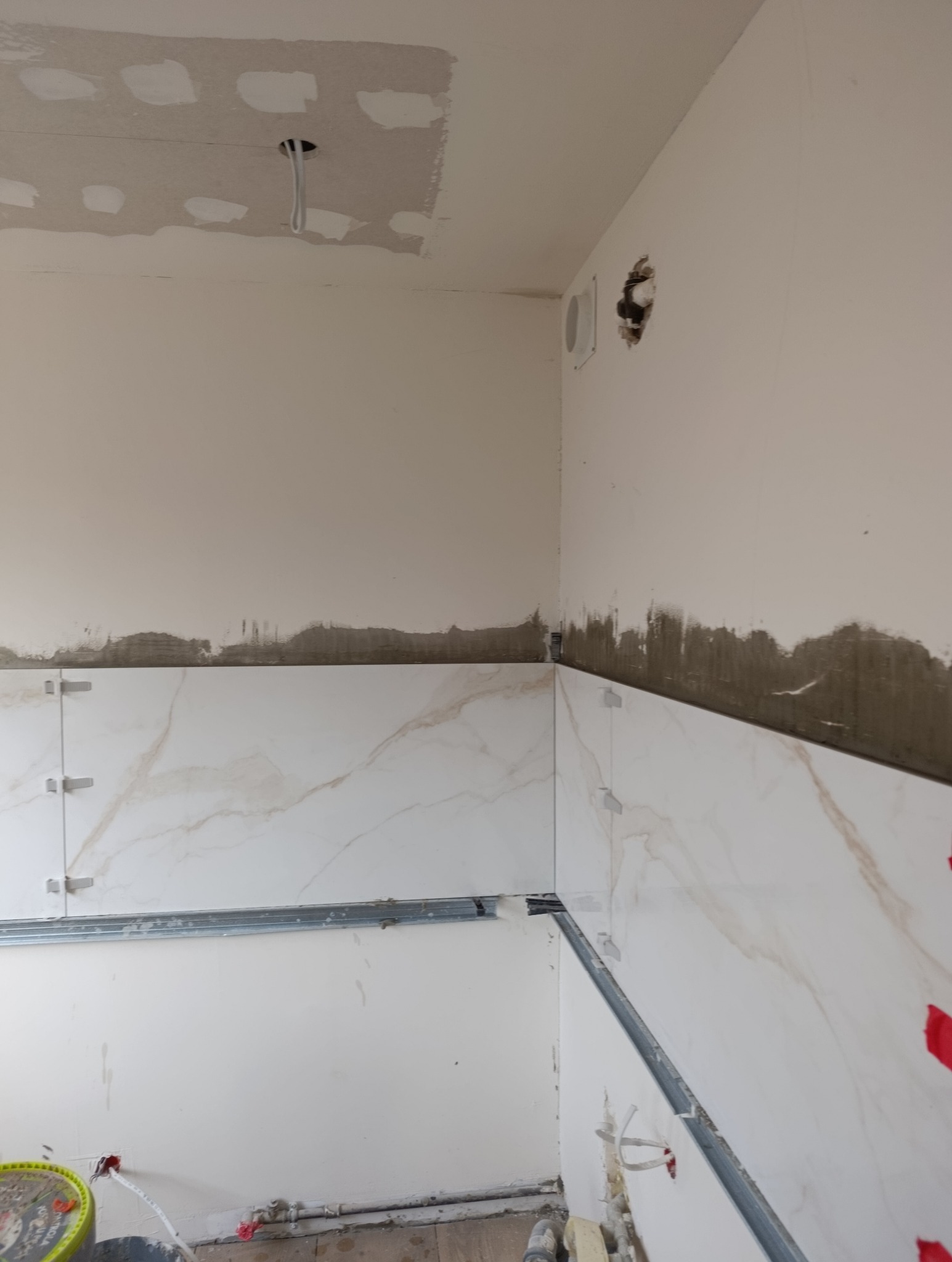 Wnętrze w trakcie prac glazurniczych: białe płytki imitujące marmur, metalowe listwy poziomujące, widoczne ślady zaprawy na ścianach i surowy sufit z instalacją.