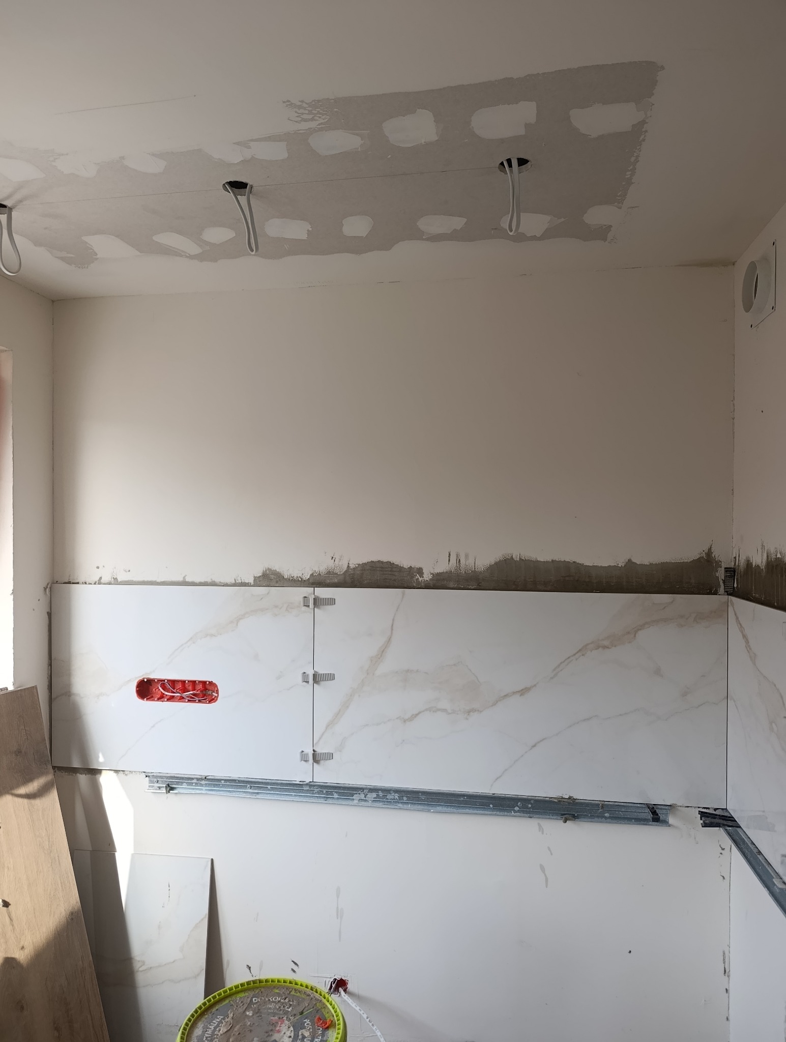 Wnętrze w trakcie remontu: białe płytki imitujące marmur na ścianie, widoczne uchwyty montażowe, puszka elektryczna i niedokończone prace na suficie.