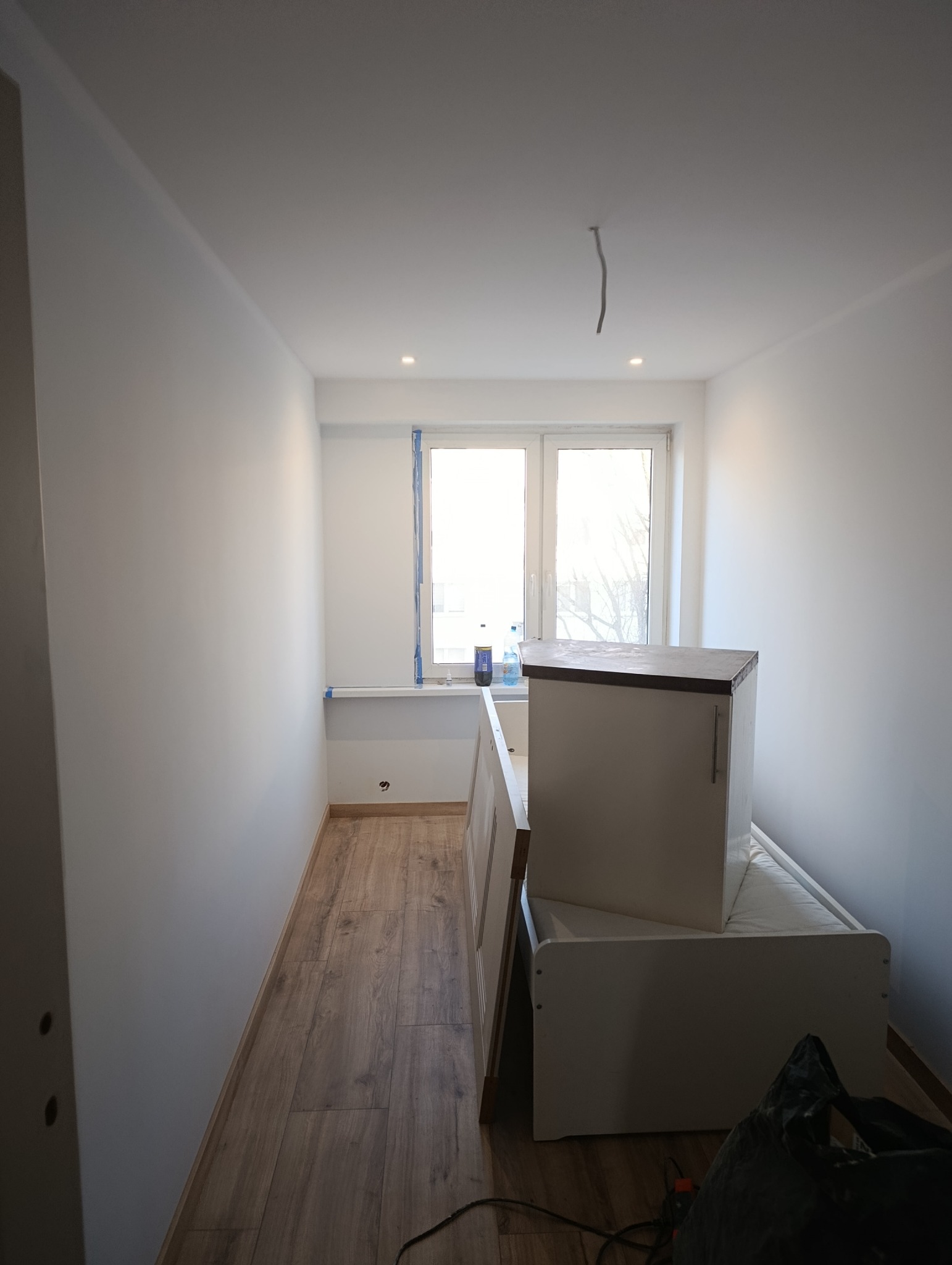 Wnętrze pokoju po malowaniu ścian na biało i ułożeniu paneli podłogowych. Widoczne okno z zabezpieczeniem taśmą malarską i meble w trakcie montażu.