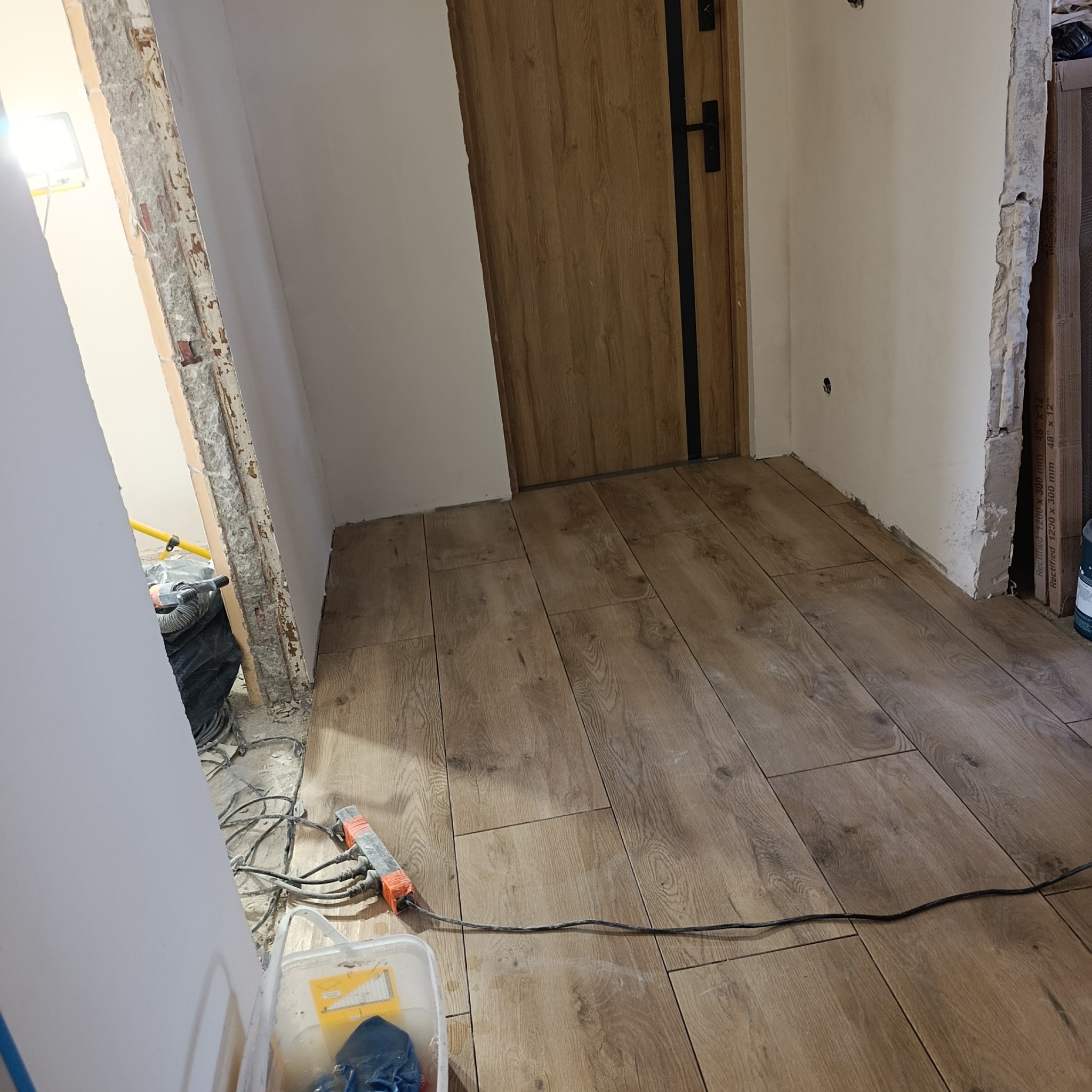 Ułożone panele podłogowe w odcieniach brązu, widoczne narzędzia pracy i fragment drzwi. Częściowo wykończone ściany w pomieszczeniu.