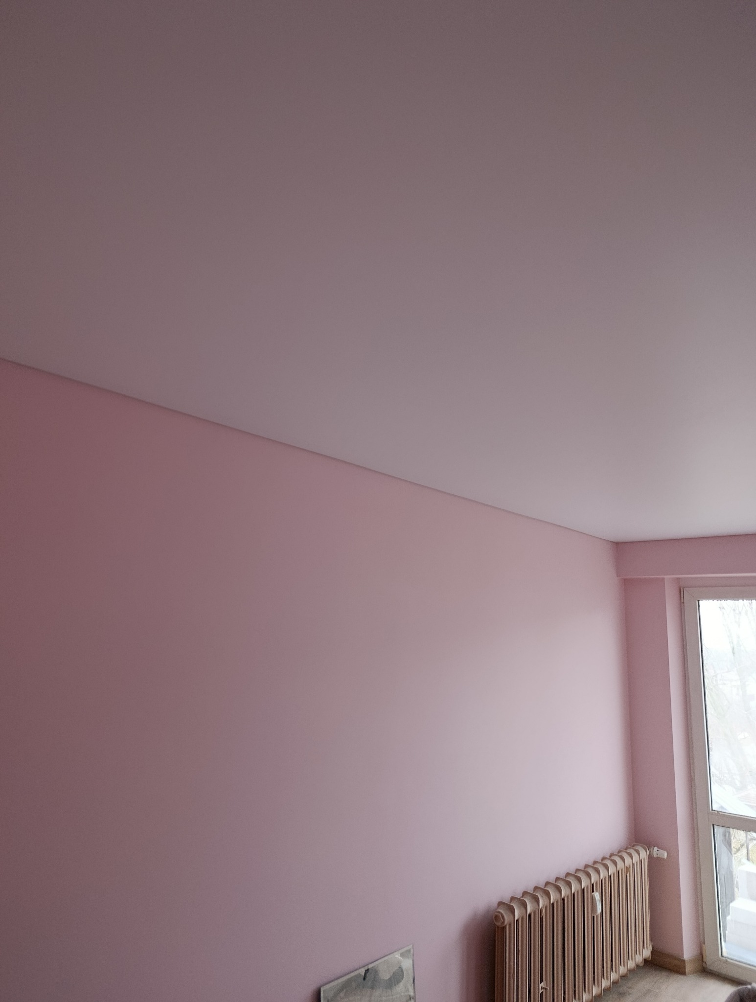 Różowa ściana po malowaniu z grzejnikiem i oknem. Widoczny fragment sufitu. Świeżo pomalowane wnętrze w pastelowych barwach.