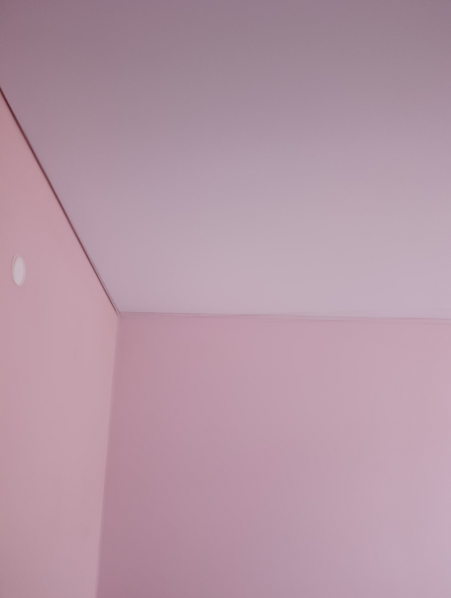 Różowe ściany i sufit w rogu pokoju, widoczna lampa sufitowa. Świeżo pomalowane powierzchnie, gładkie wykończenie. Minimalistyczny design wnętrza.