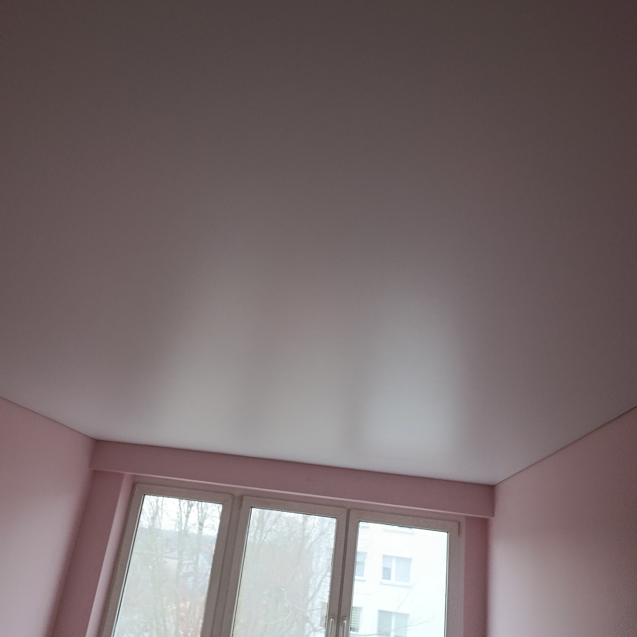 Biały sufit napinany w pokoju z różowymi ścianami i oknem. Widok z dołu, ukazujący gładką powierzchnię sufitu i fragmenty ścian oraz okna.