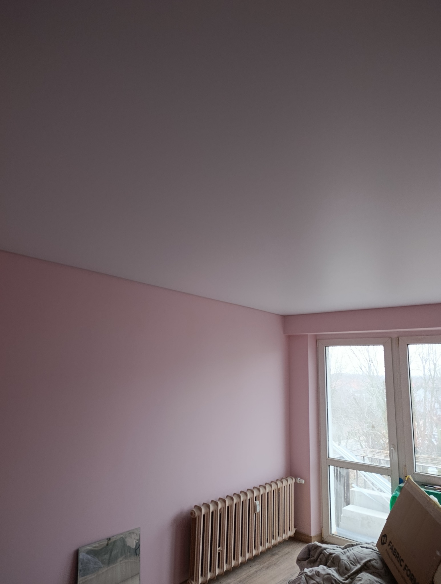 Różowe ściany po malowaniu w pokoju z grzejnikiem i oknem balkonowym. Widoczny fragment podłogi i karton. Świeżo pomalowane ściany w pastelowym odcieniu.