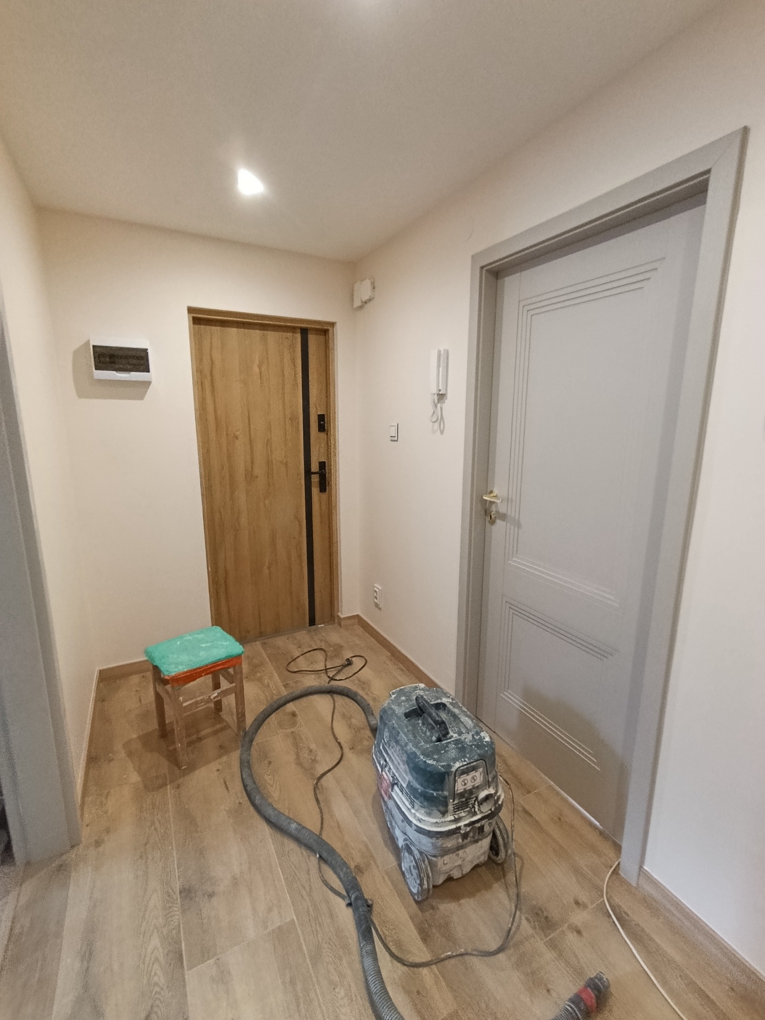 Korytarz po remoncie: ułożone panele podłogowe, świeżo pomalowane ściany, zamontowane drzwi. Widoczny odkurzacz przemysłowy i stołek malarski.