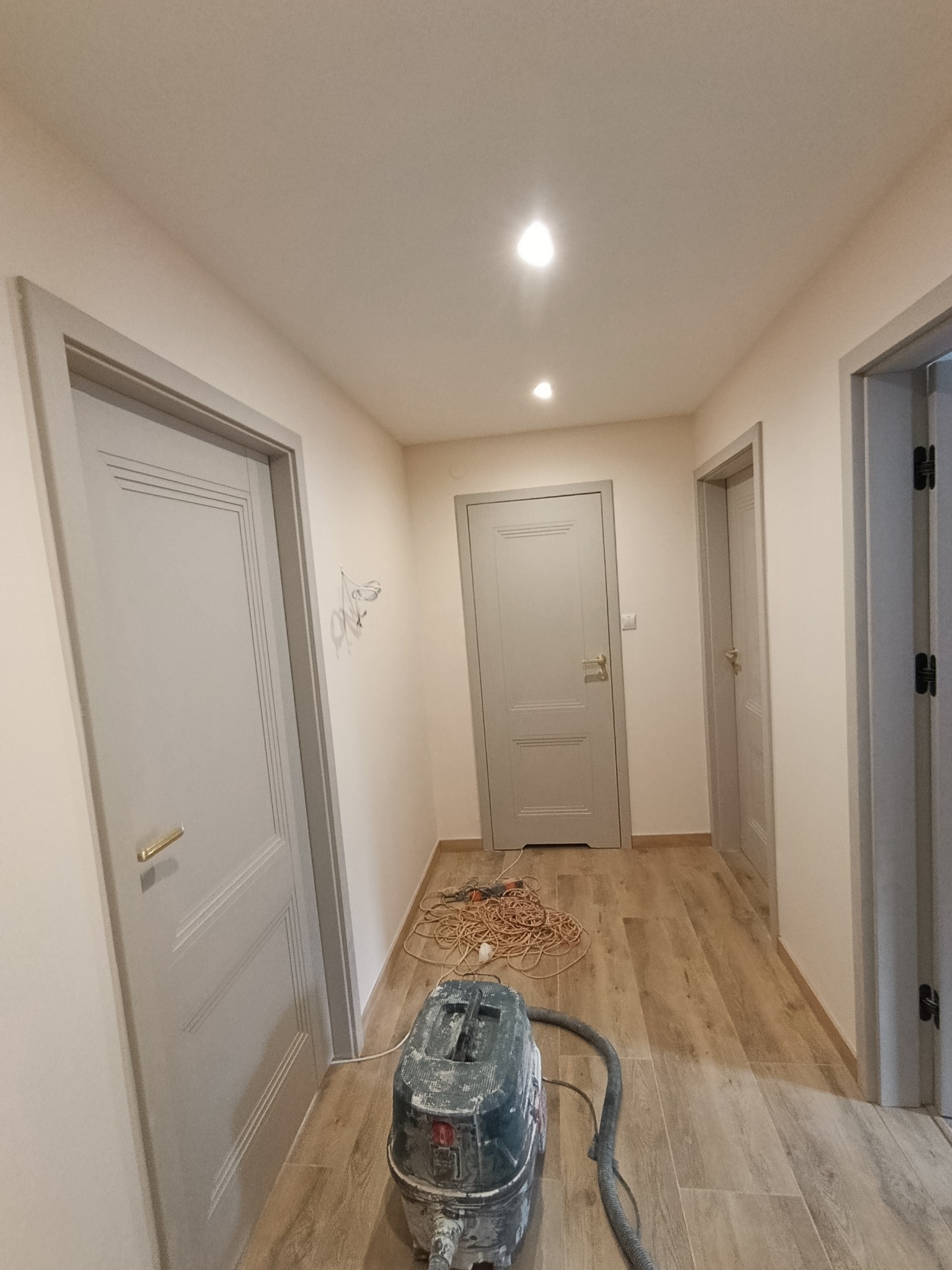Korytarz po remoncie z trzema szarymi drzwiami, drewnopodobną podłogą i odkurzaczem przemysłowym. Widoczne ślady po instalacji na ścianie.