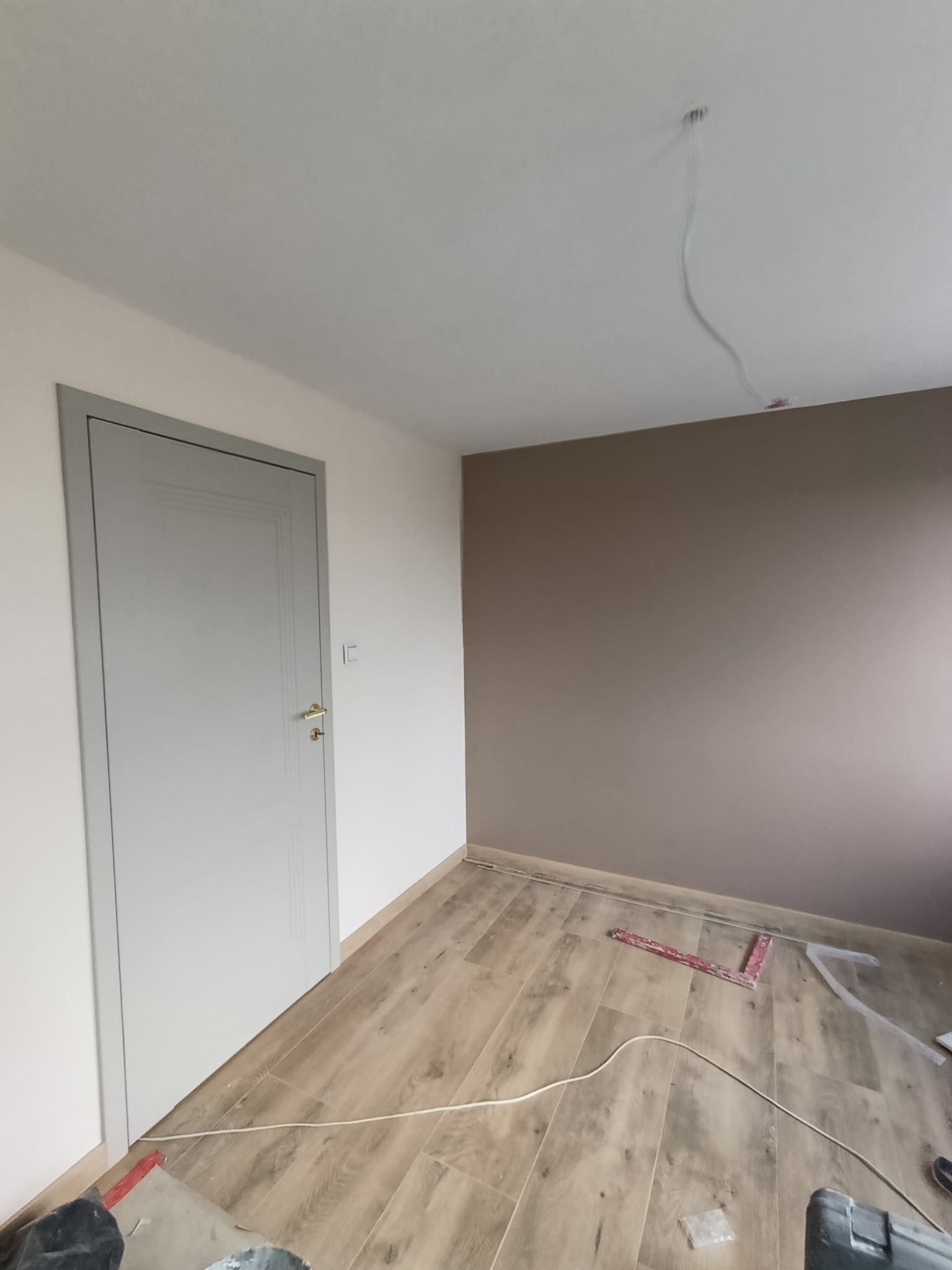 Jasne panele podłogowe w pokoju z szarą ościeżnicą i drzwiami. Na ścianie widoczny kabel. Częściowo pomalowana ściana na brązowo. Widoczne narzędzia.