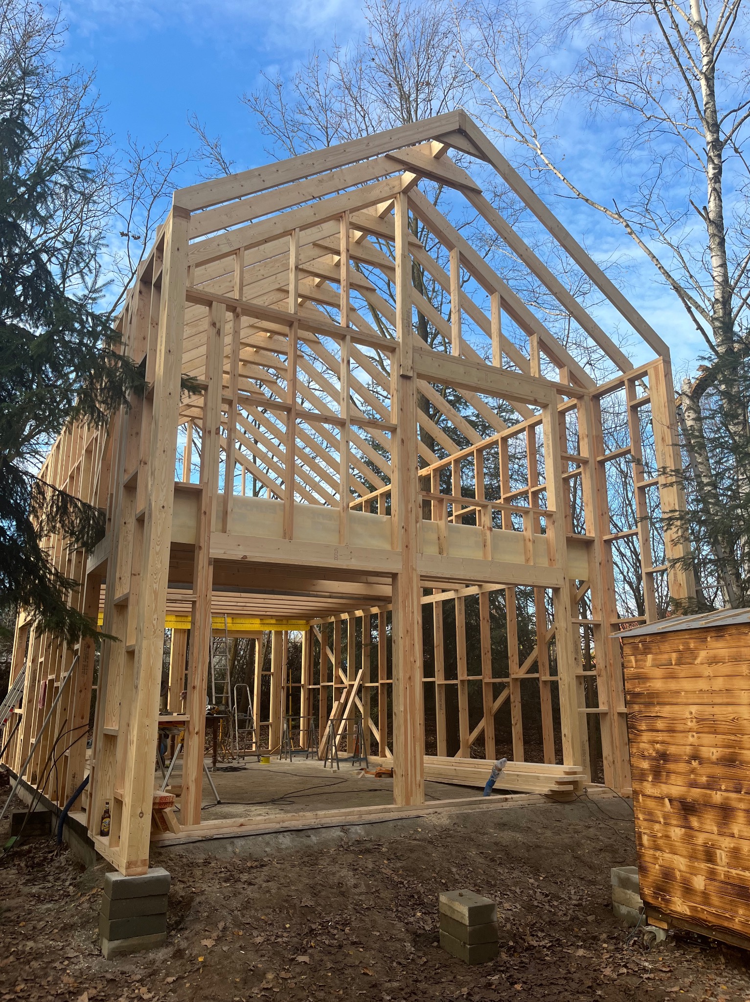 Konstrukcja szkieletowa domu w trakcie budowy, widok z zewnątrz, drewniane elementy na fundamencie, błękitne niebo w tle, drzewa bez liści.