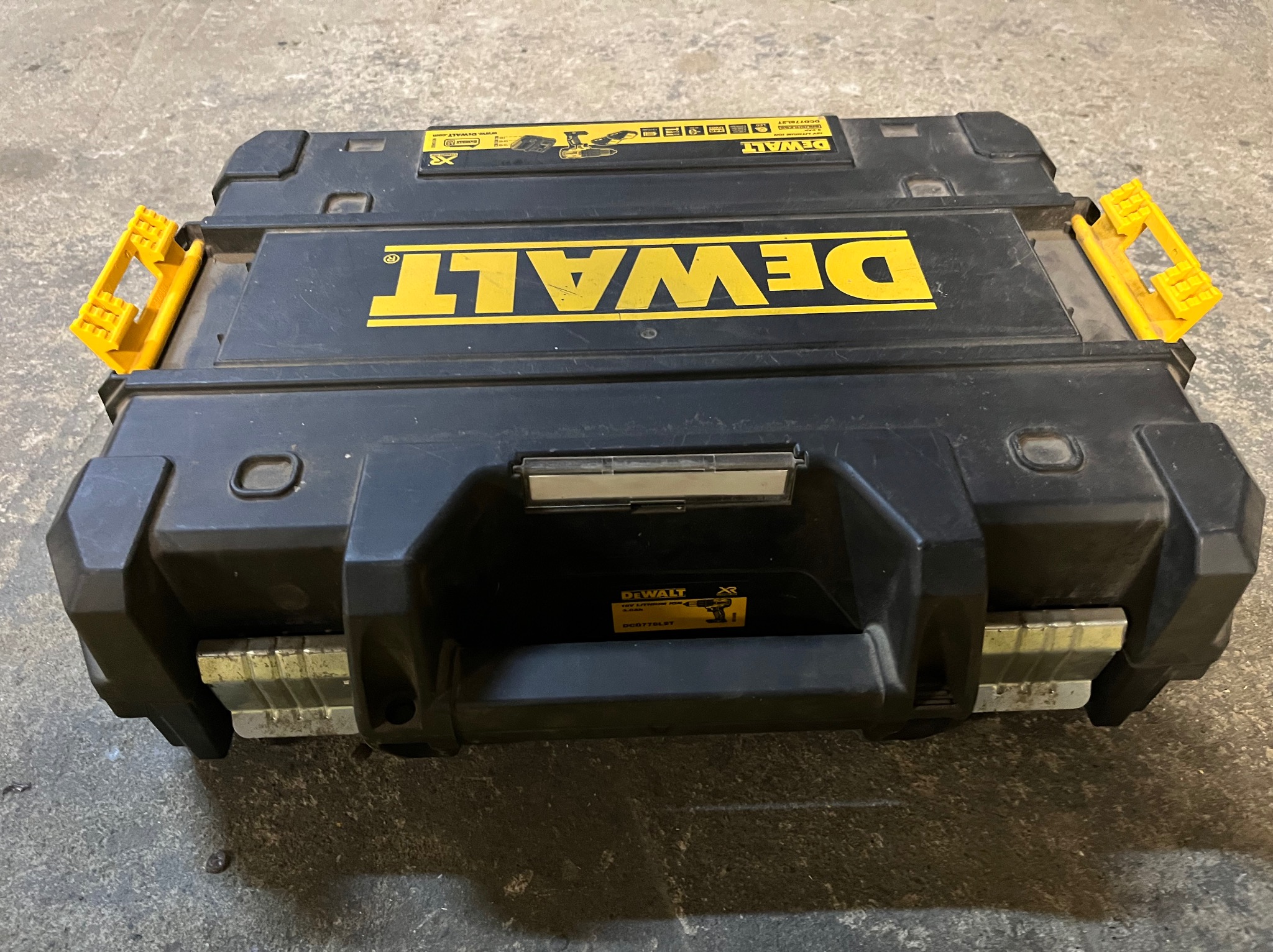 Czarna skrzynka narzędziowa DeWalt z żółtymi akcentami, leżąca na szarej, betonowej podłodze. Widoczne logo i oznaczenia modelu narzędzia.